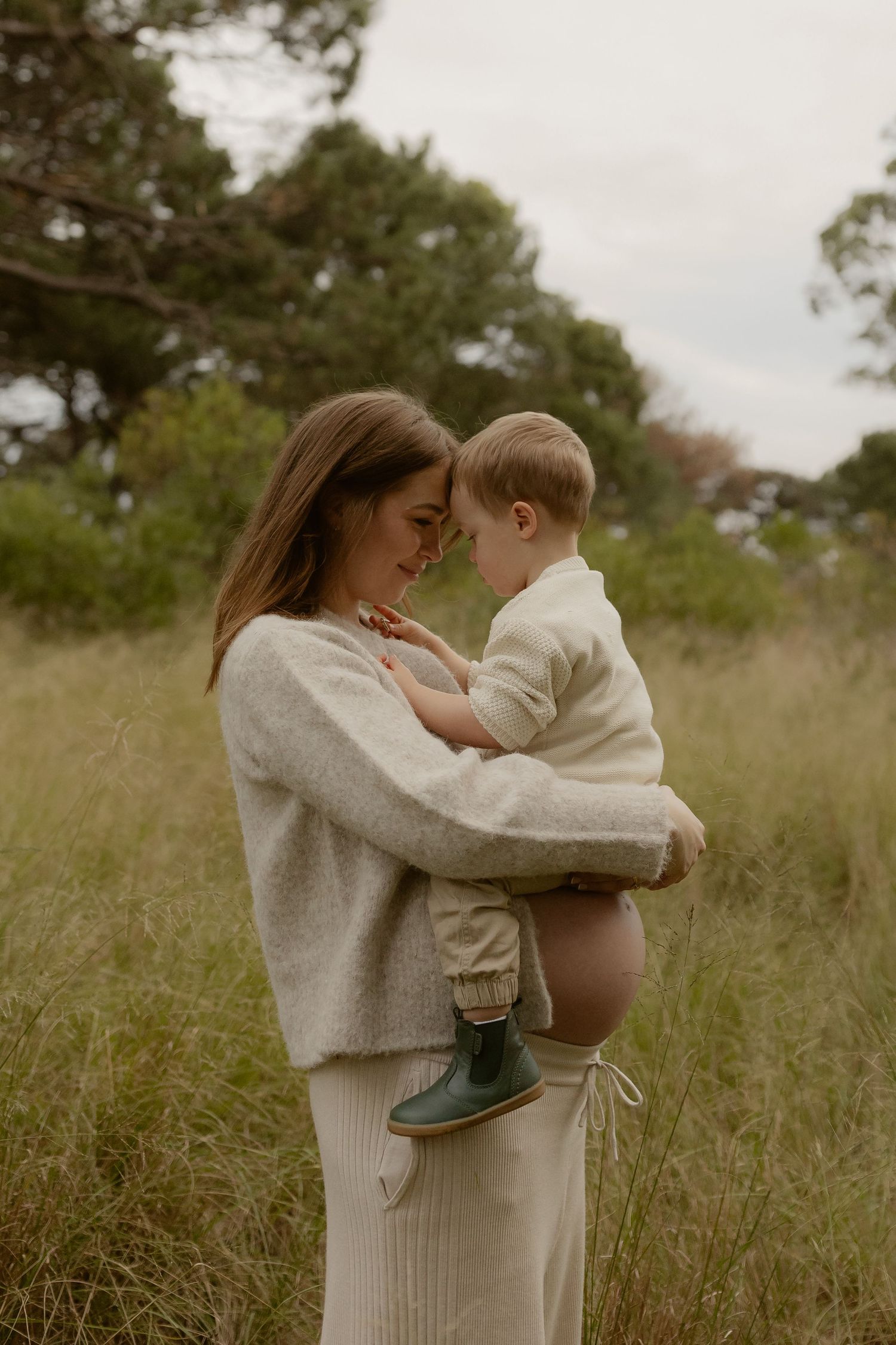 Pregnant woman in beige sweater holding toddler nose-to-nose in grassy field.