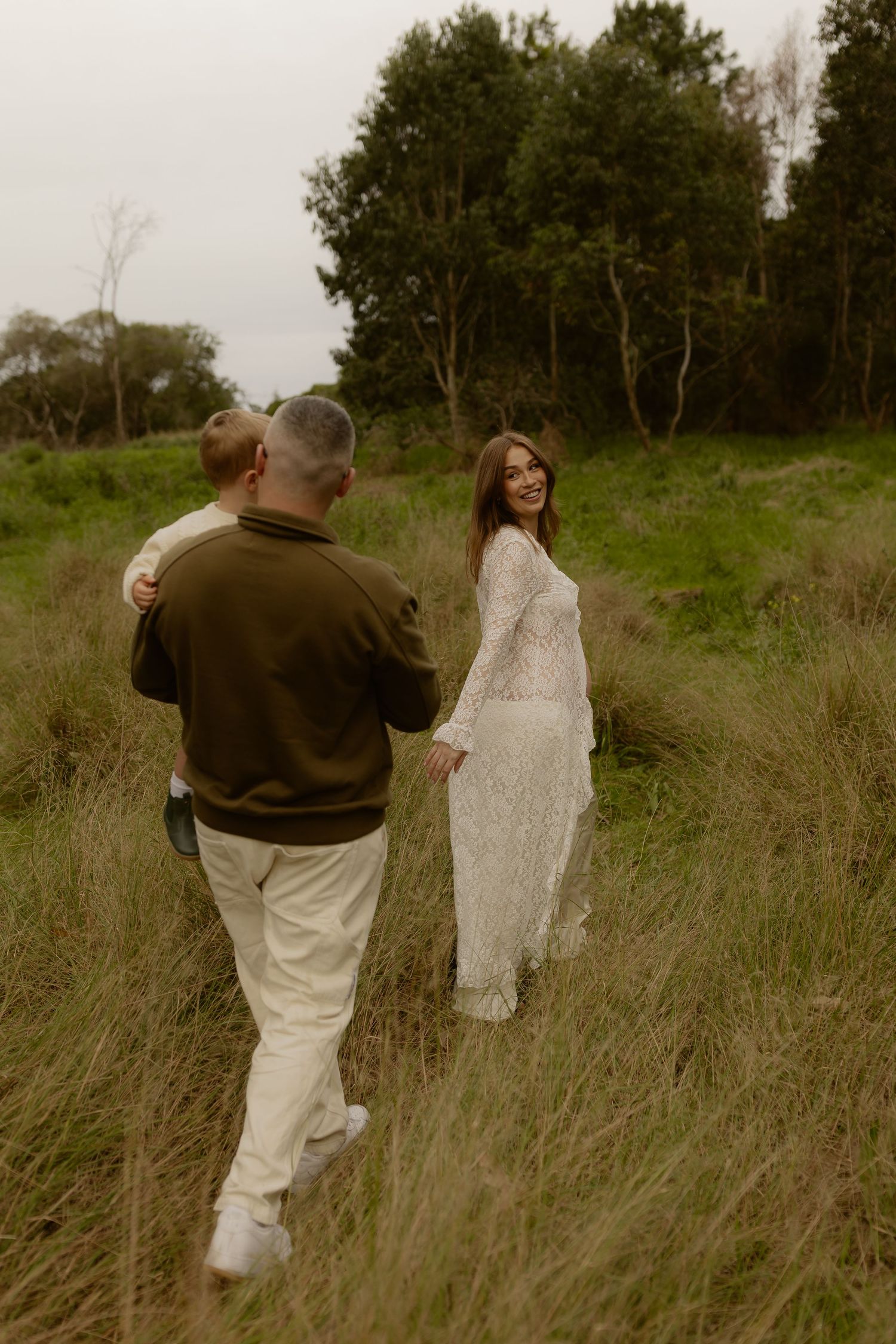 Pregnant woman in lace dress smiling back at partner and child walking through tall grass.