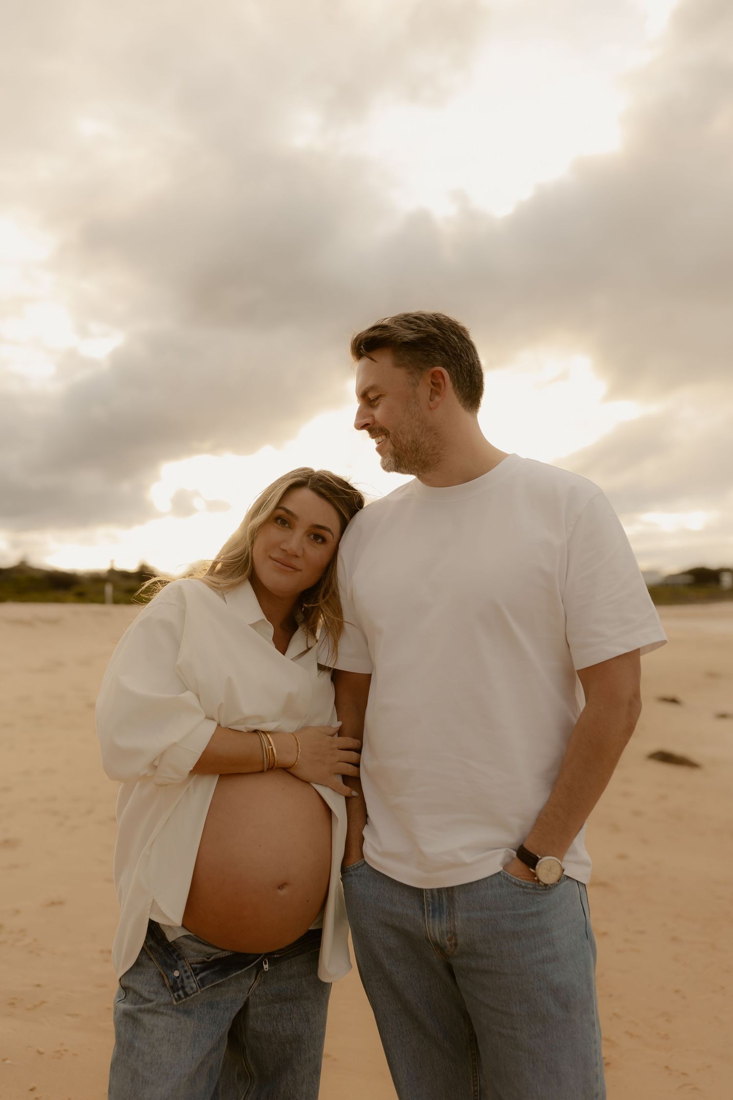 Smiling pregnant woman leans on partner on sandy beach at sunset, both in white tops and jeans.