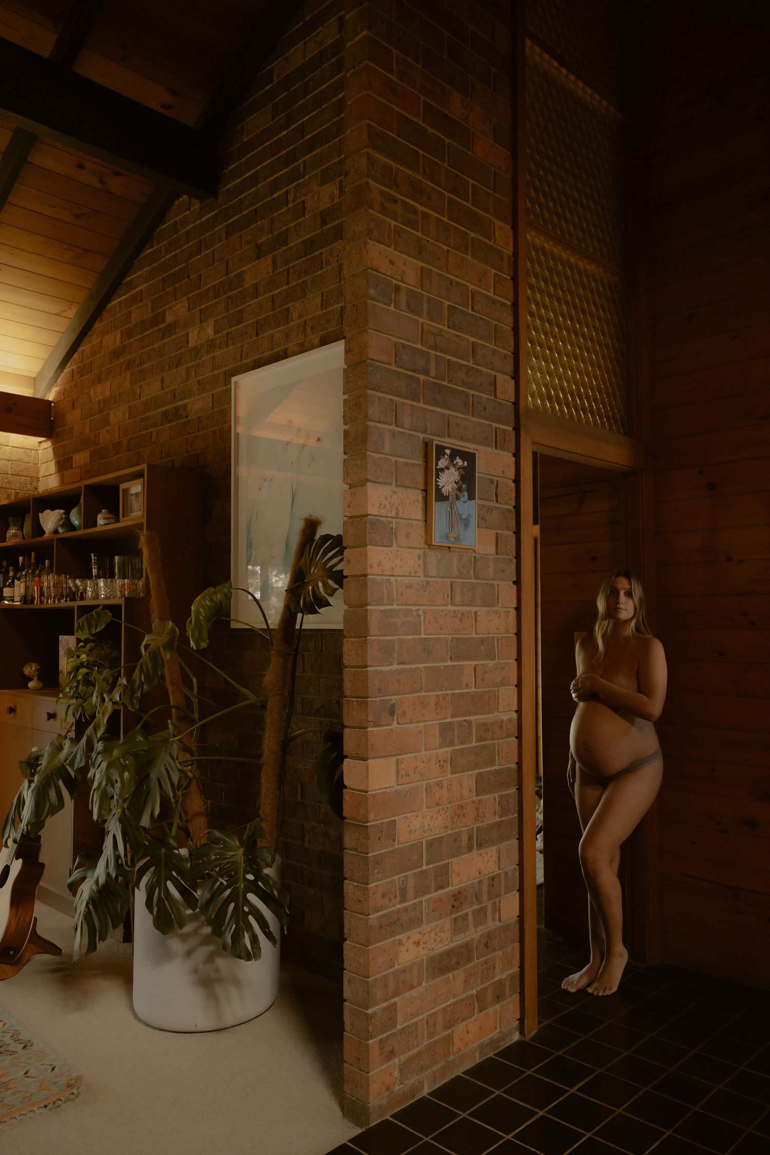 Pregnant woman posing nude beside a brick wall in a warm, rustic interior with wood ceilings and lush plants.