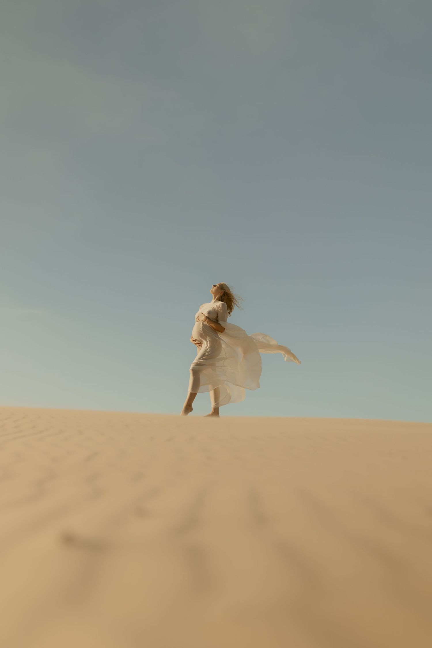 Woman in white dress walking up a sand dune against a clear blue sky in the desert.