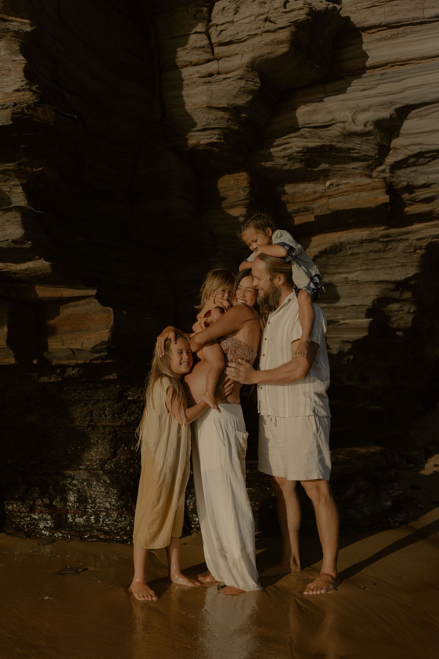 Family of four poses barefoot near rocky cliffs on a beach, parents embracing children in warm golden light.