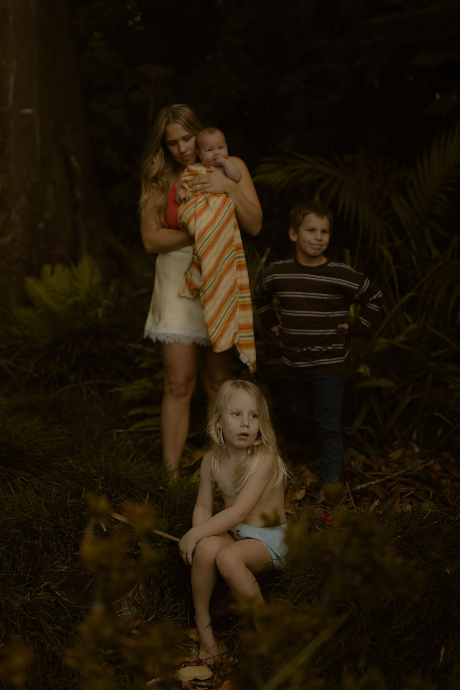 A mother holding a newborn in a striped blanket with two children in a lush forest setting.