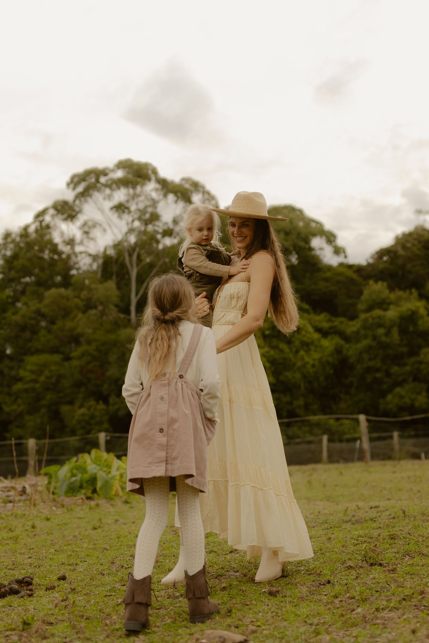 A woman in a straw hat and cream dress holds a toddler while a girl watches on a farm.