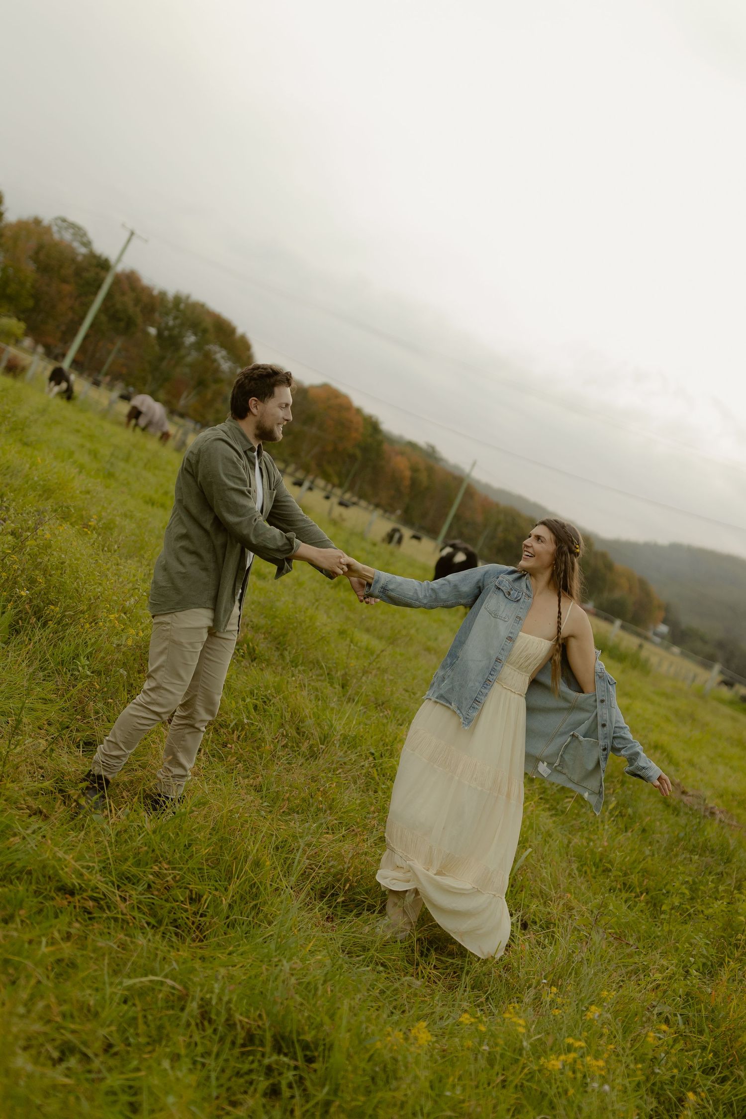 A couple holds hands and dances in a lush green field on a cloudy autumn day.