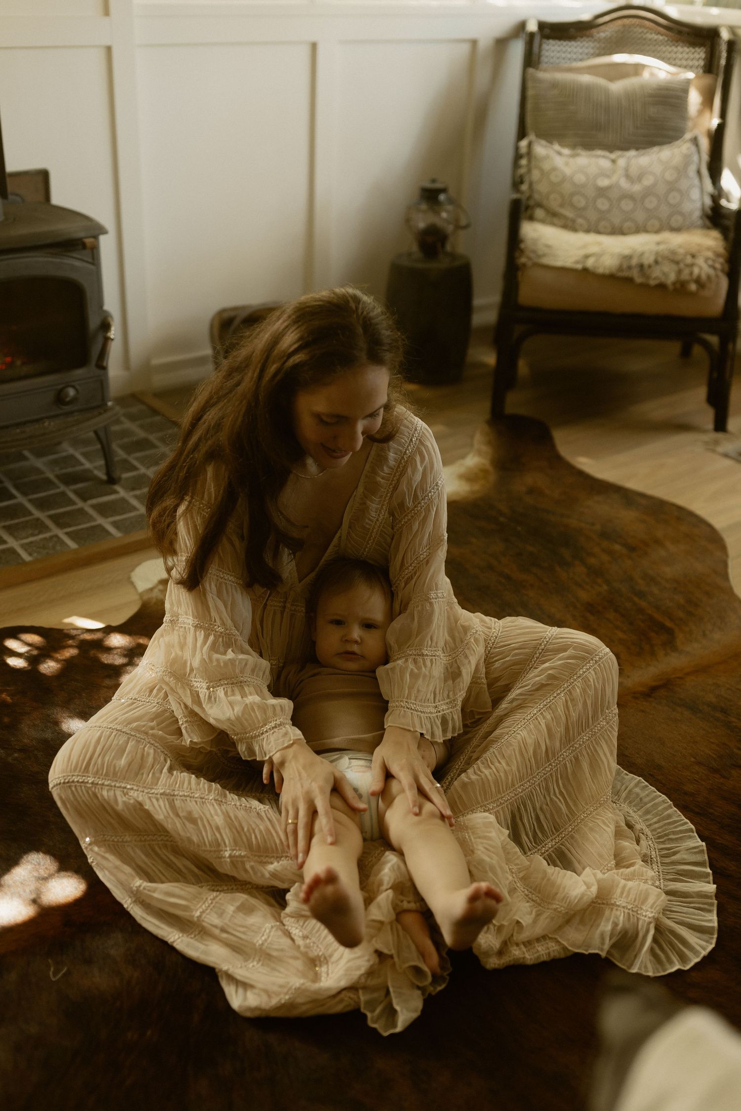 A dreamy sunlit scene shows a mother and baby sitting on a hardwood floor in white flowing dresses.
