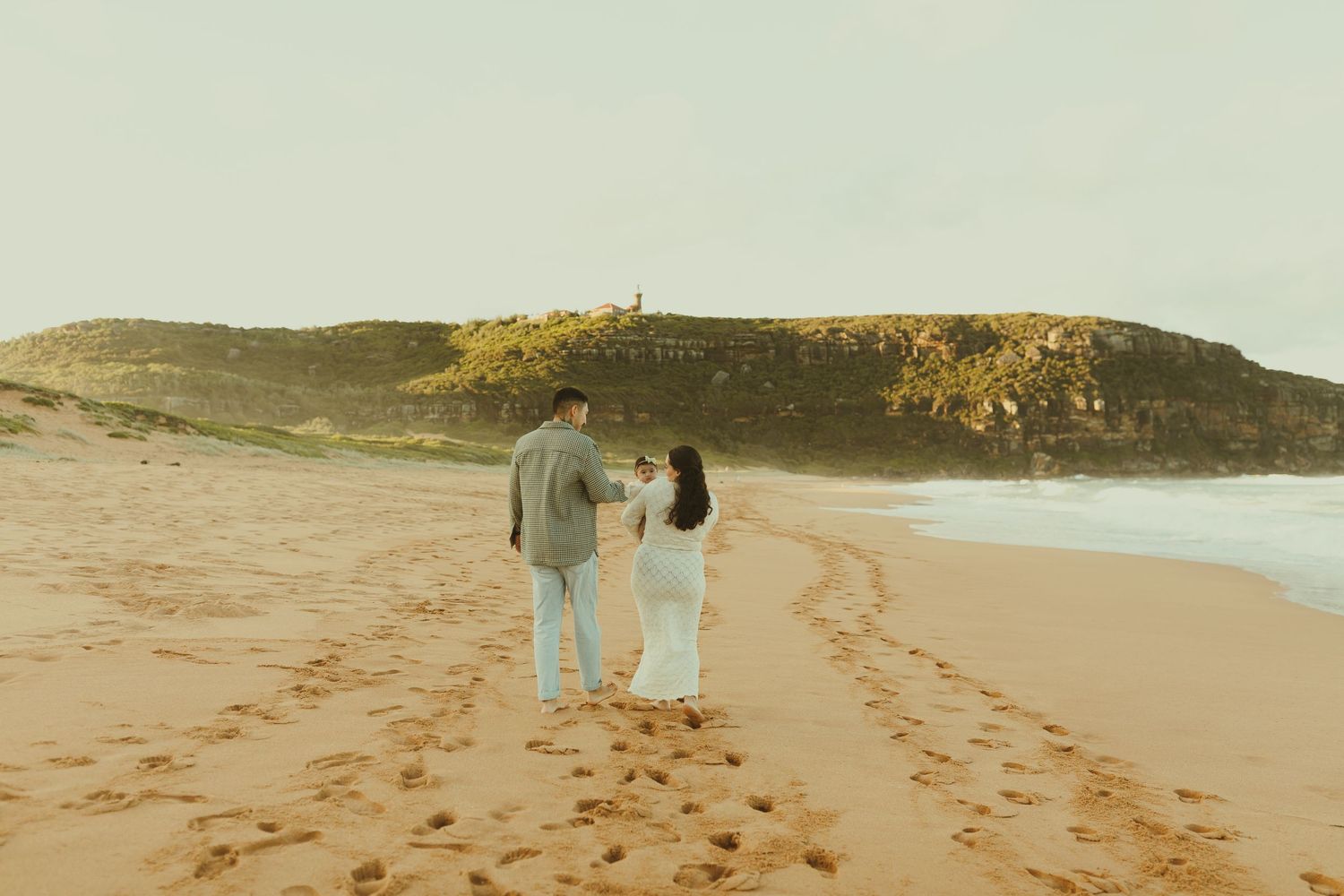 Pair walking along a scenic beach shoreline with lighthouse and cliffs visible in the distance on a hazy day.