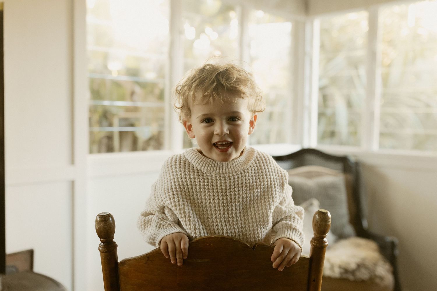 Joyful toddler sitting in wooden chair wearing white knit sweater in sunlit room.