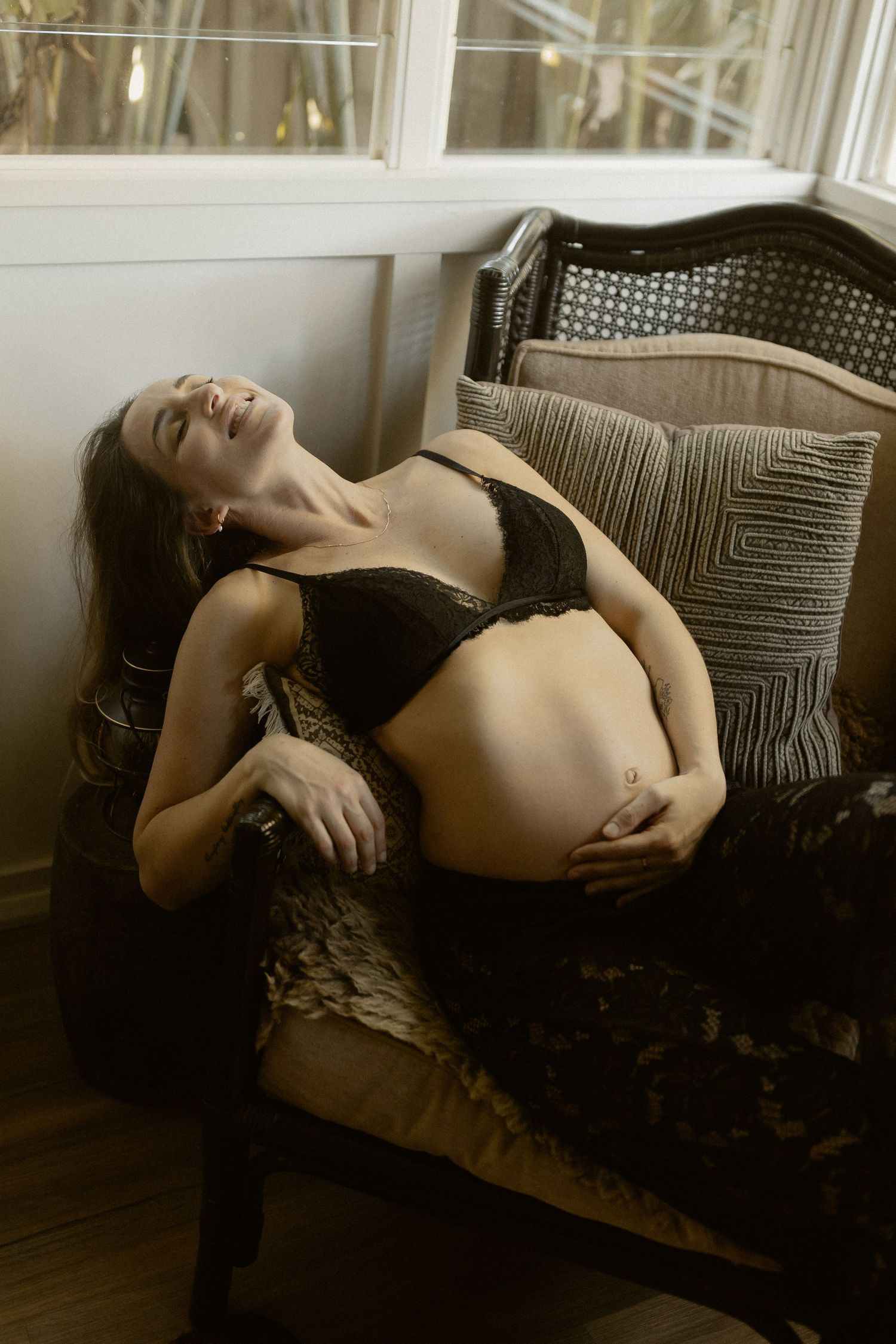 Intimate maternity portrait showing pregnant woman relaxing on antique chaise in natural window light.