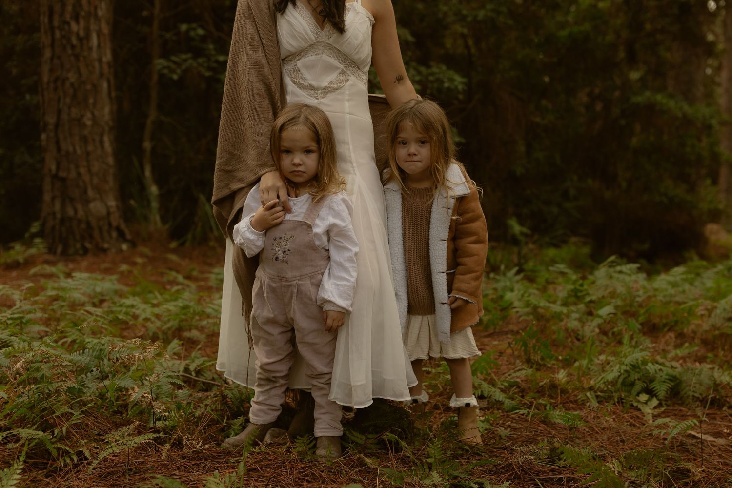 Two young children stand beside a woman in a white dress in a lush forest setting, wearing neutral toned outfits.