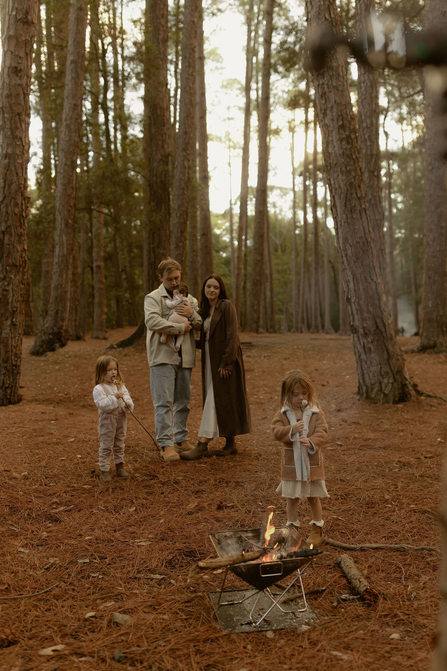Family gathered around a campfire in a pine forest, with two children and parents enjoying an outdoor autumn evening.