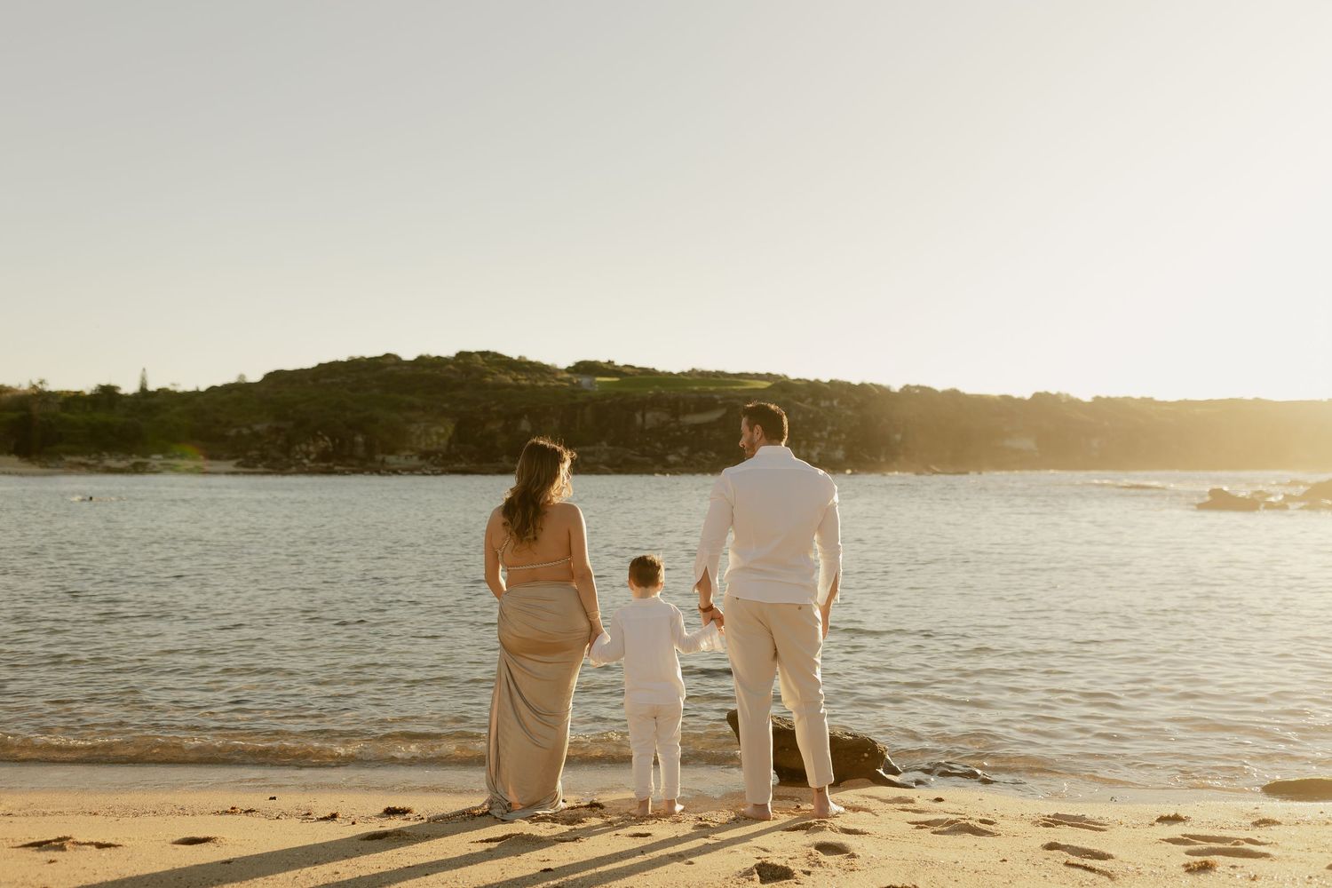Family of three holding hands on a sandy beach at golden sunset, with lush green hills and calm water in the background.
