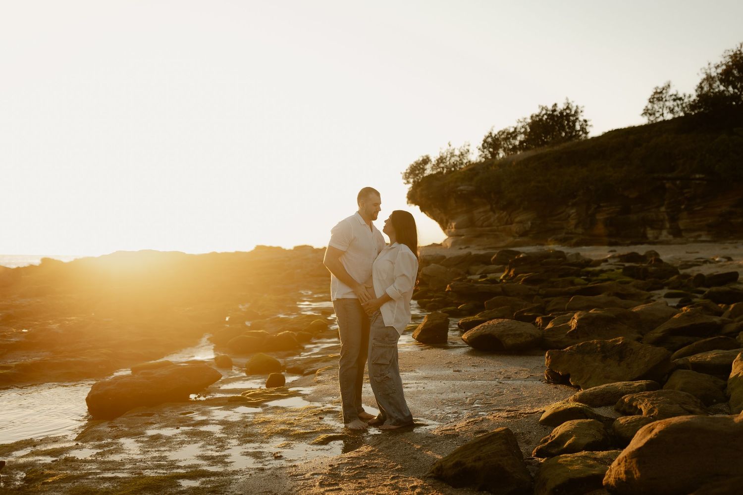 Couple embracing on rocky beach at golden sunset with glowing cliffs in background.
