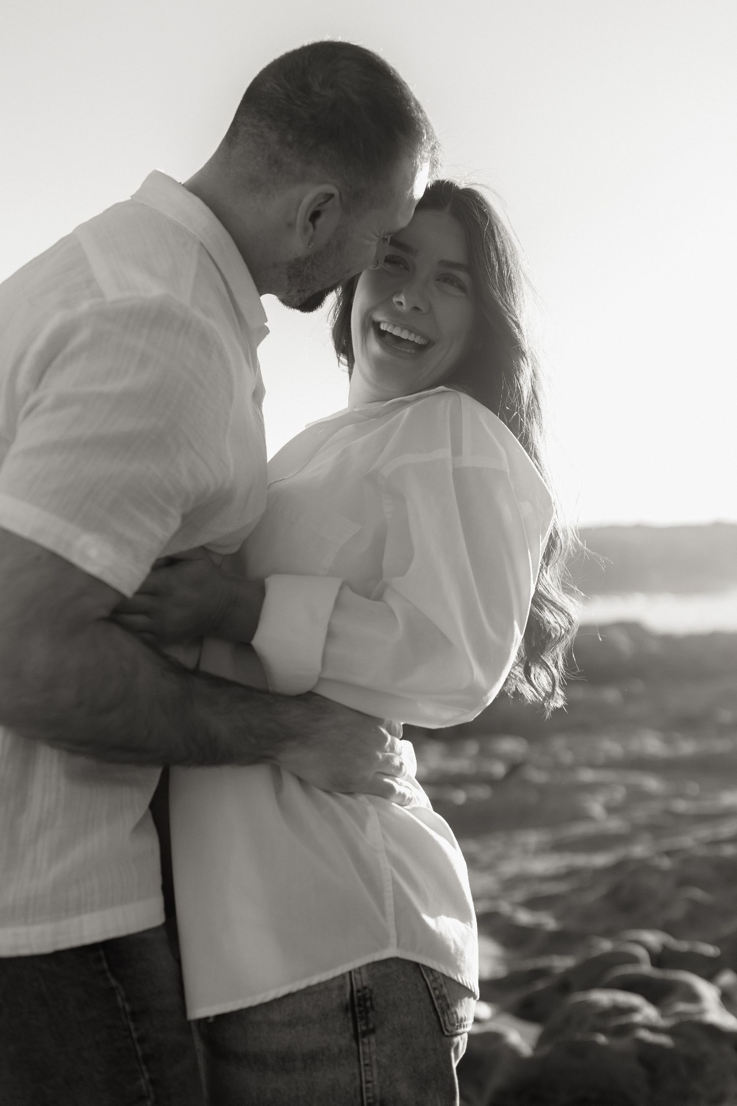 Black and white photo of couple embracing and laughing on rocky beach at sunset.