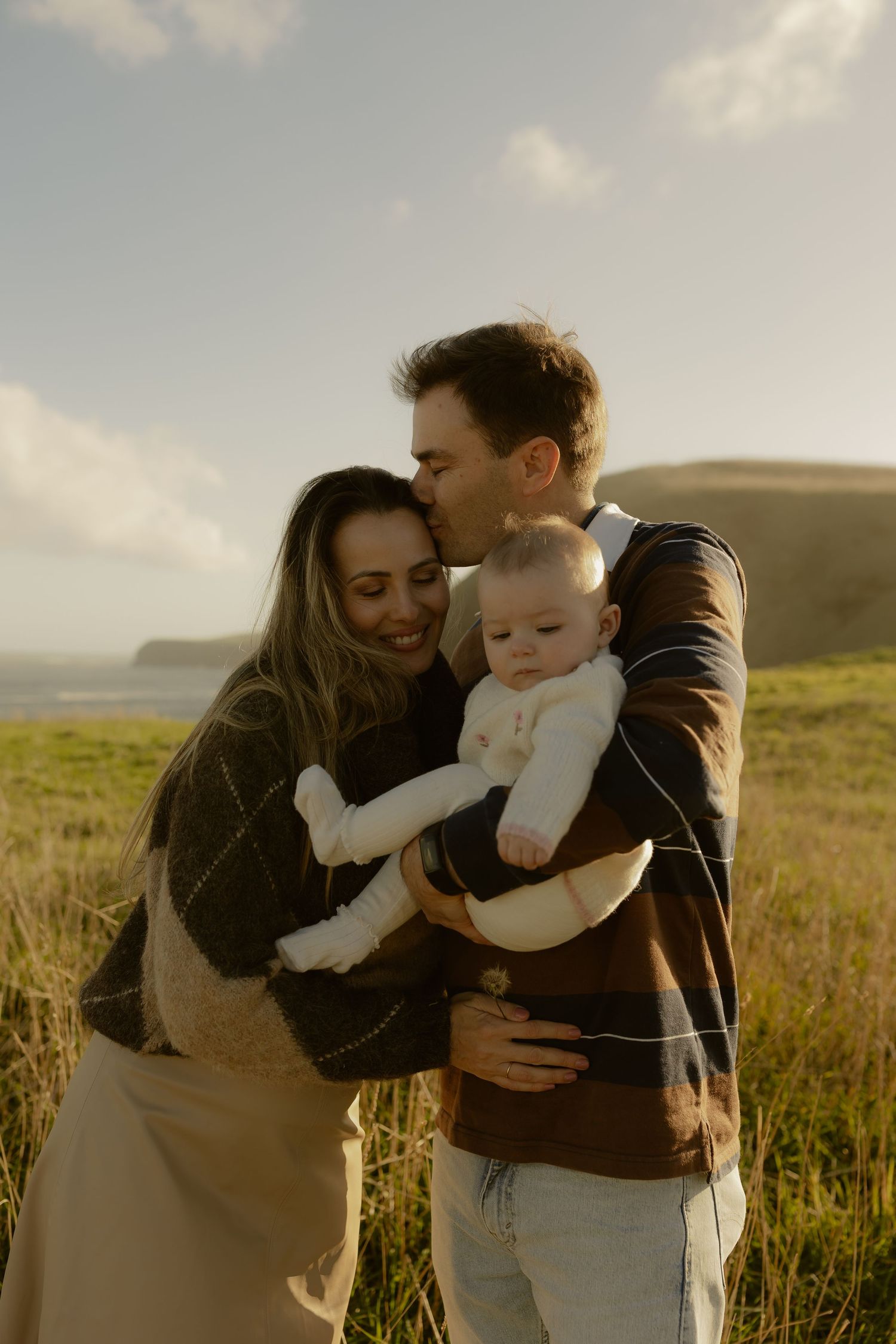 A couple stands in a golden field, man kissing woman's head as they hold their smiling baby in white.