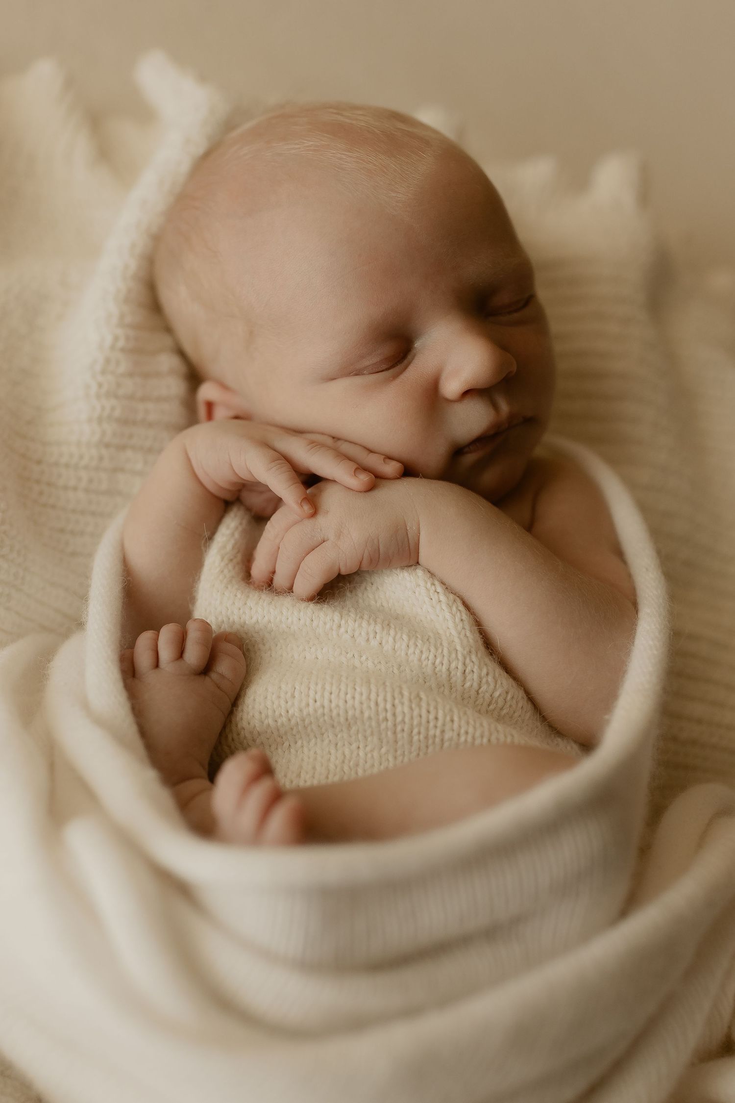 A peaceful sleeping newborn baby wrapped in soft cream-colored knit blankets during a professional portrait session.