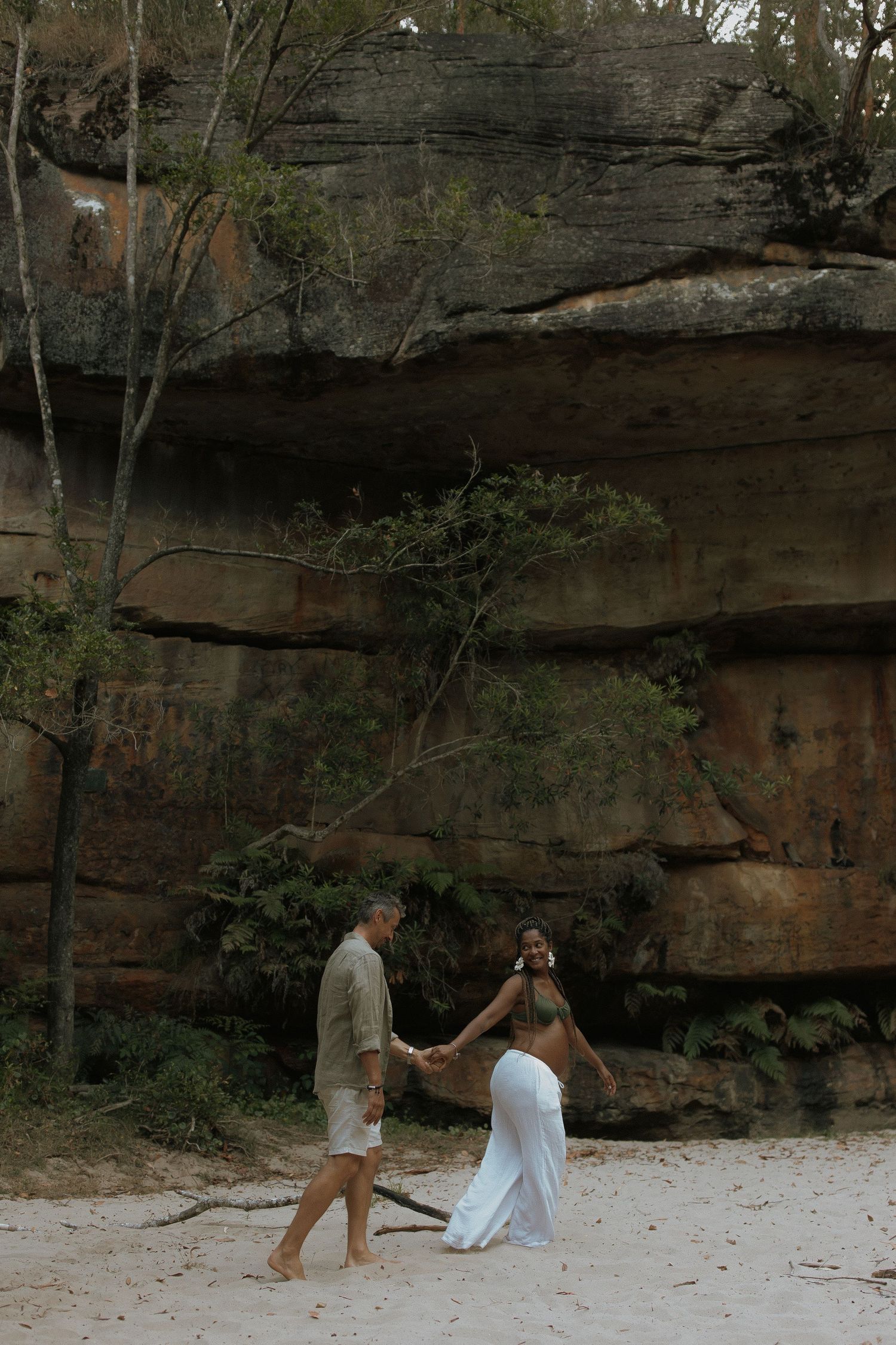 A couple in white outfits walks along a sandy path beneath towering rock cliffs in a natural outdoor setting.