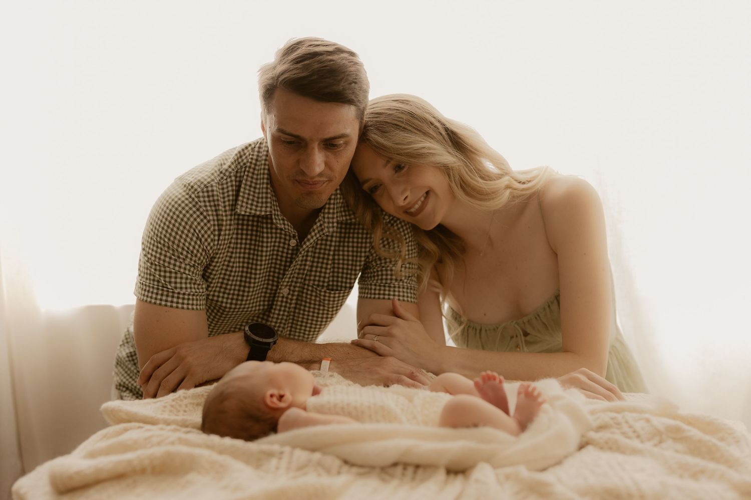 Parents lovingly gazing at their newborn baby lying on soft beige bedding in warm natural light.