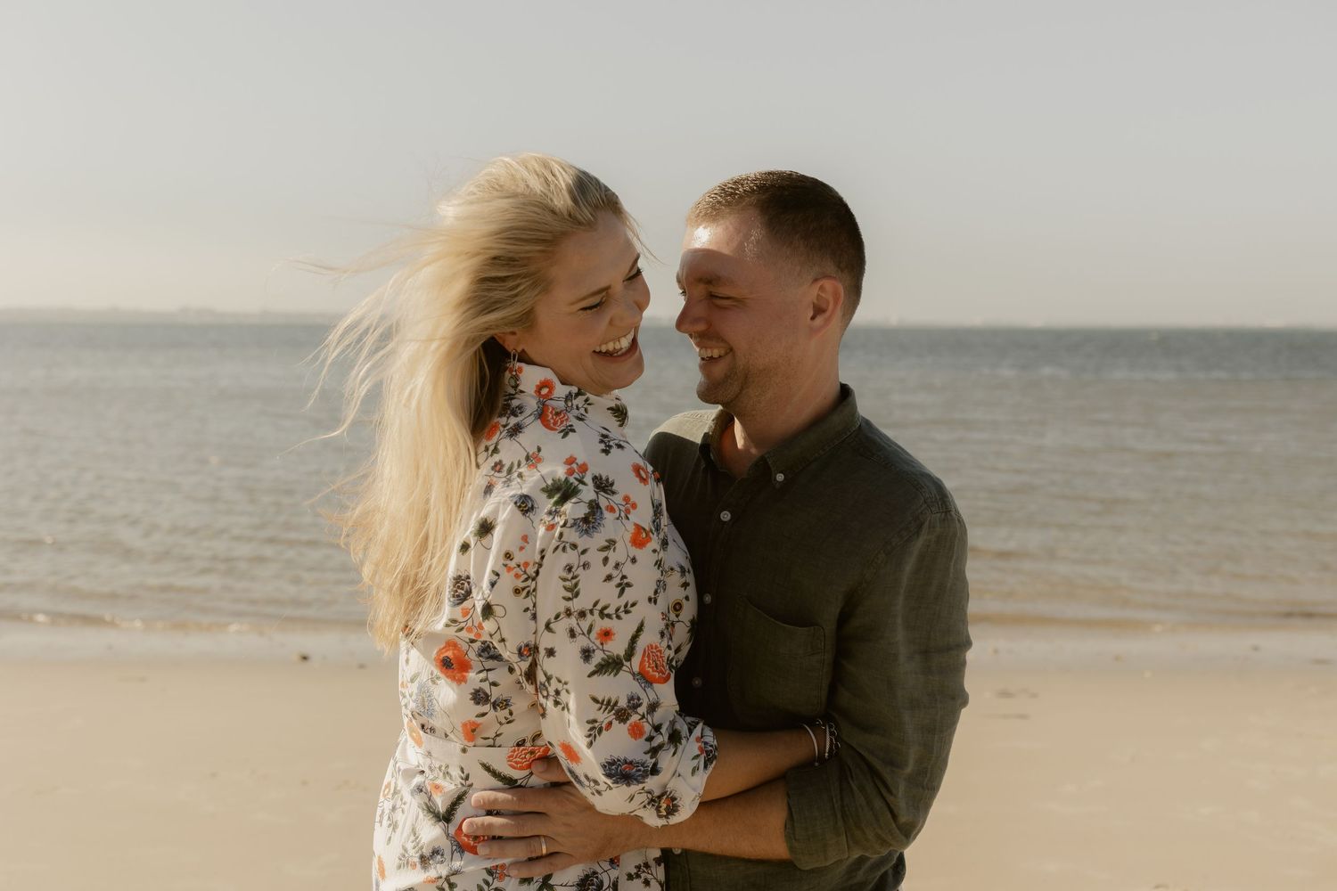 A couple embraces and laughs together during a romantic photoshoot on a windy beach at sunset.