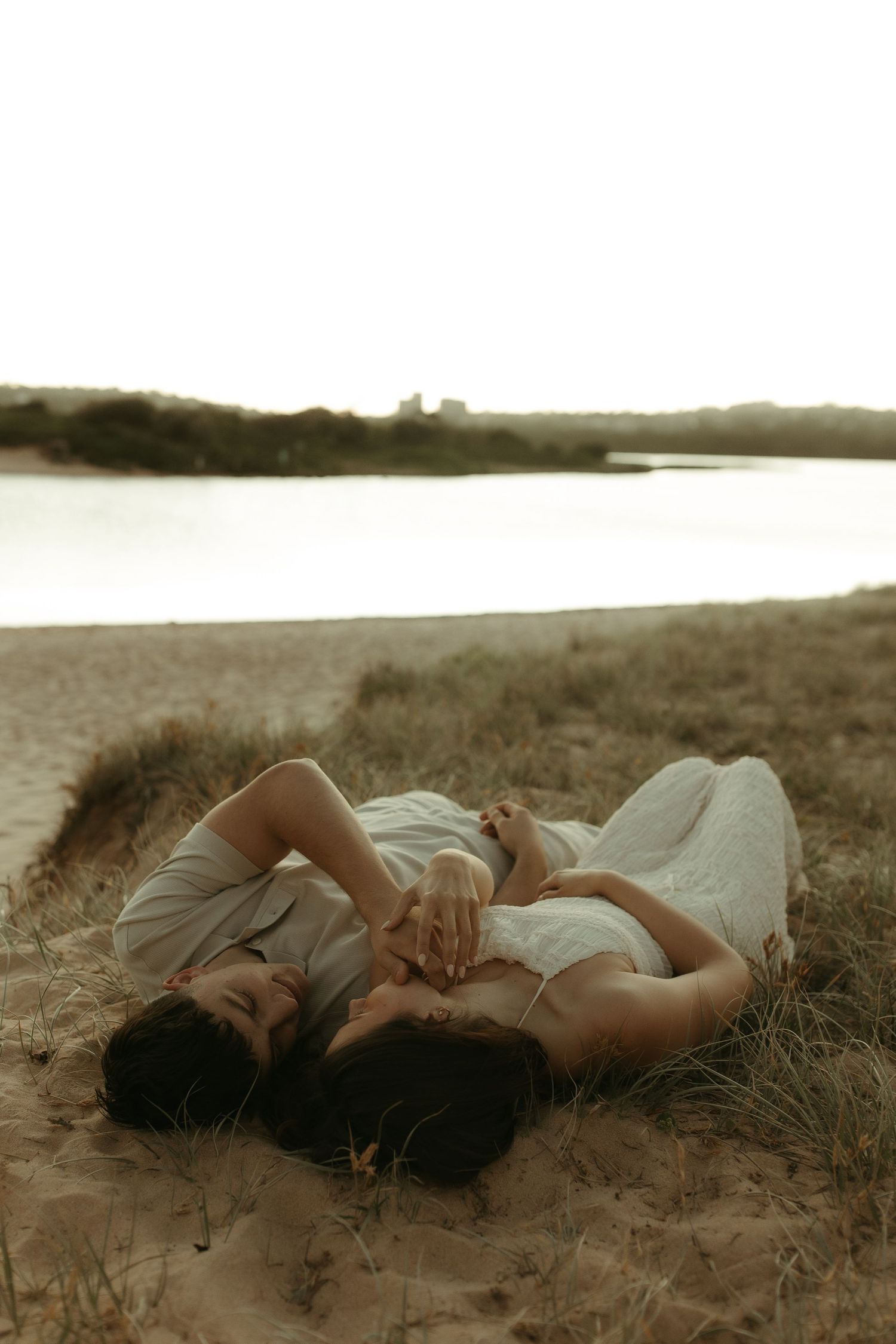 Two people in light clothing lie together on sandy ground near a tranquil lake at dusk.