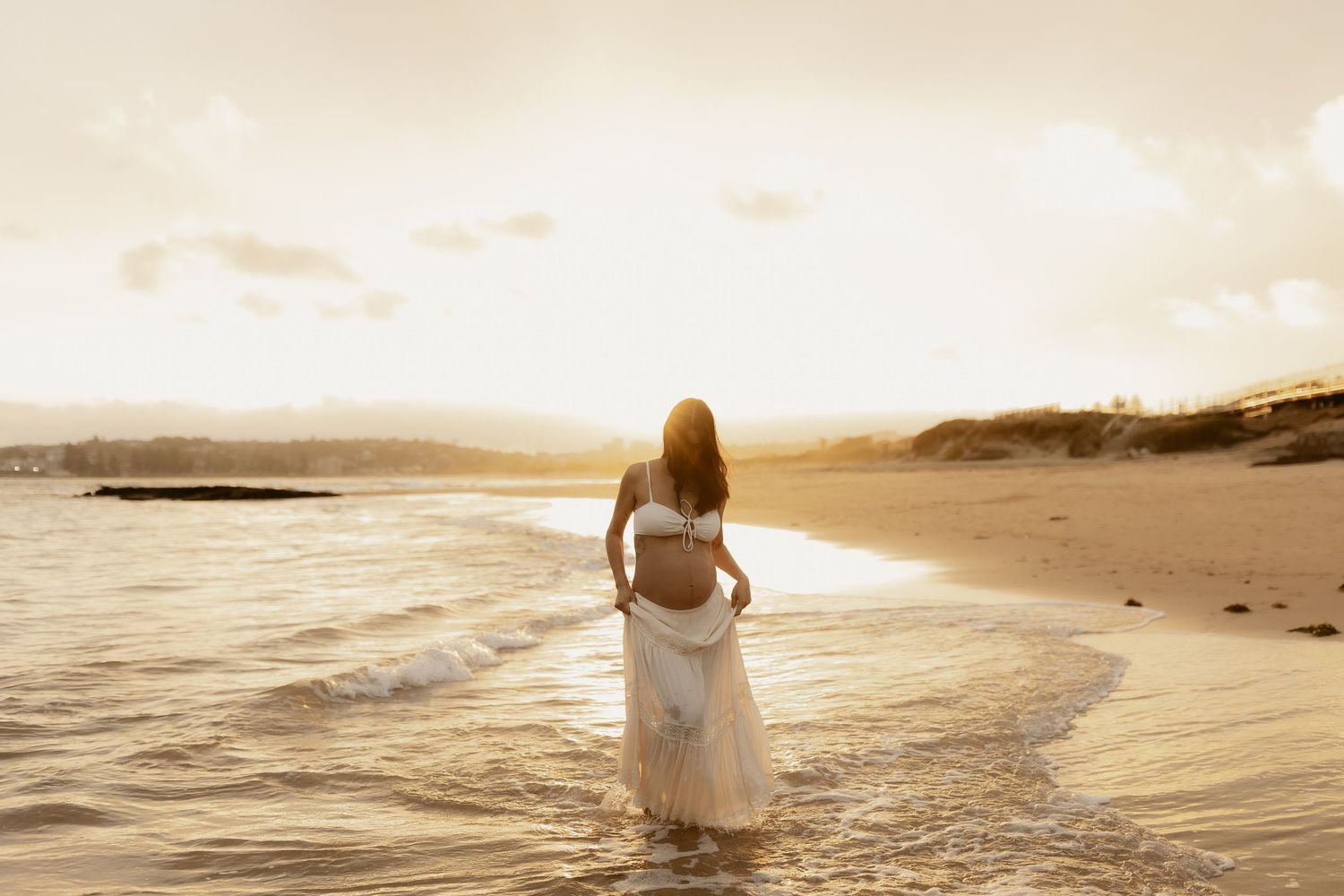 Person in white dress walking along serene beach shoreline during sunset with waves lapping at their feet.