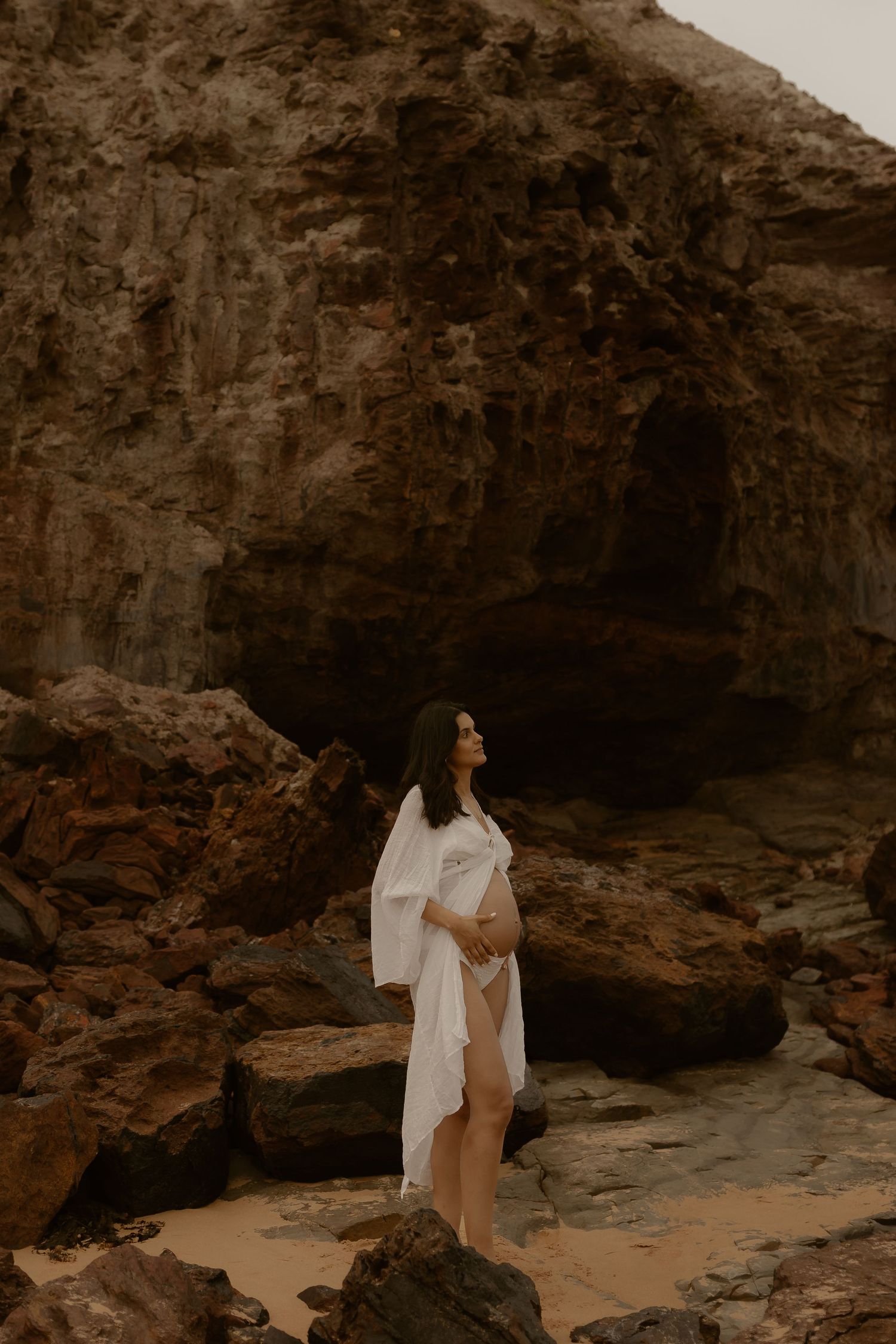 A woman in white stands contemplatively on rocky coastal cliffs near a cave entrance.