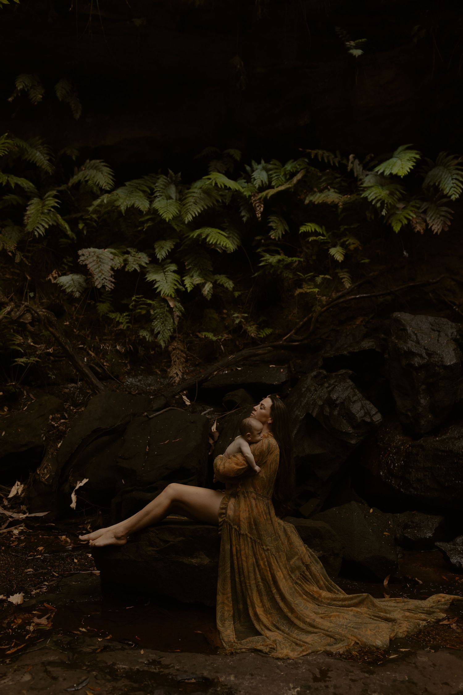 A person in a vintage brown dress reclines against dark tree roots in a moody forest setting.