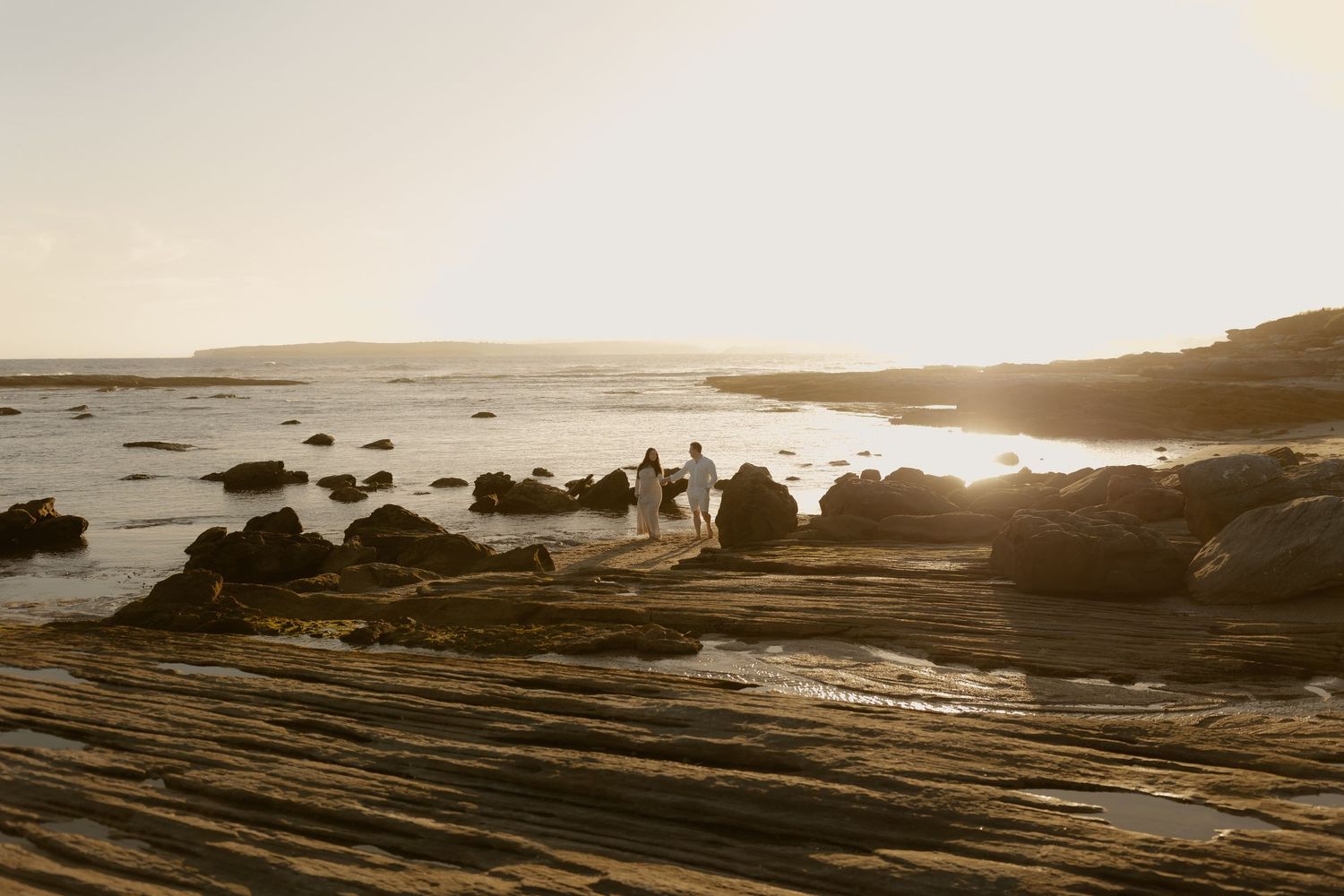 A golden sunset illuminates a rocky coastal beach landscape with silhouettes against the glowing horizon.