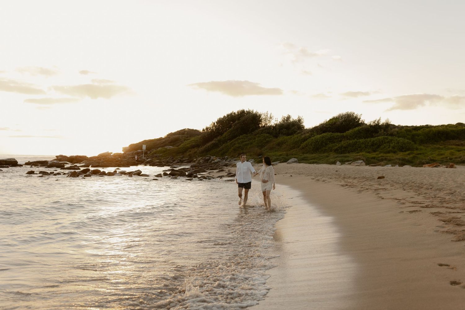 Two people walk along a scenic beach shoreline at sunset with waves rolling onto the sand.