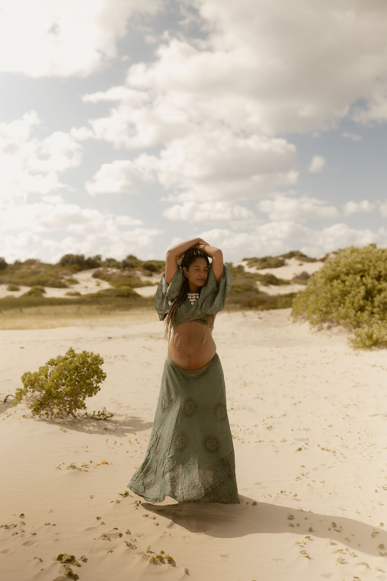 Person in olive green dress sitting among coastal sand dunes under cloudy sky.
