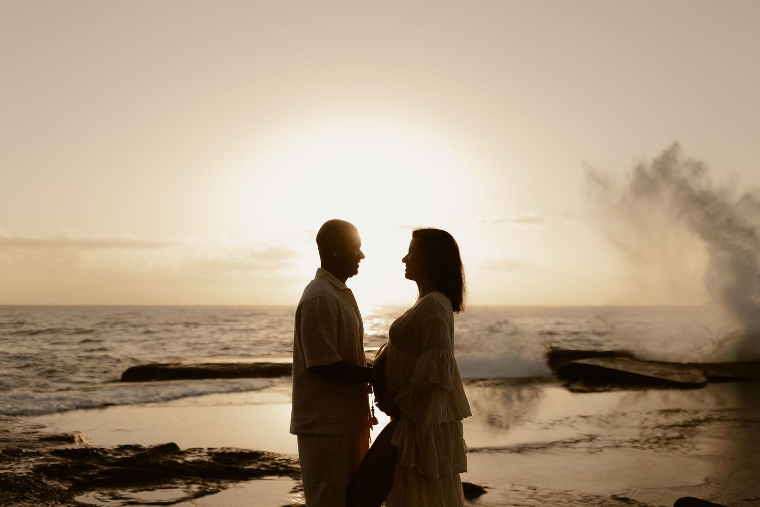 Silhouetted couple stands together on beach at sunset with ocean waves and rocks in background.