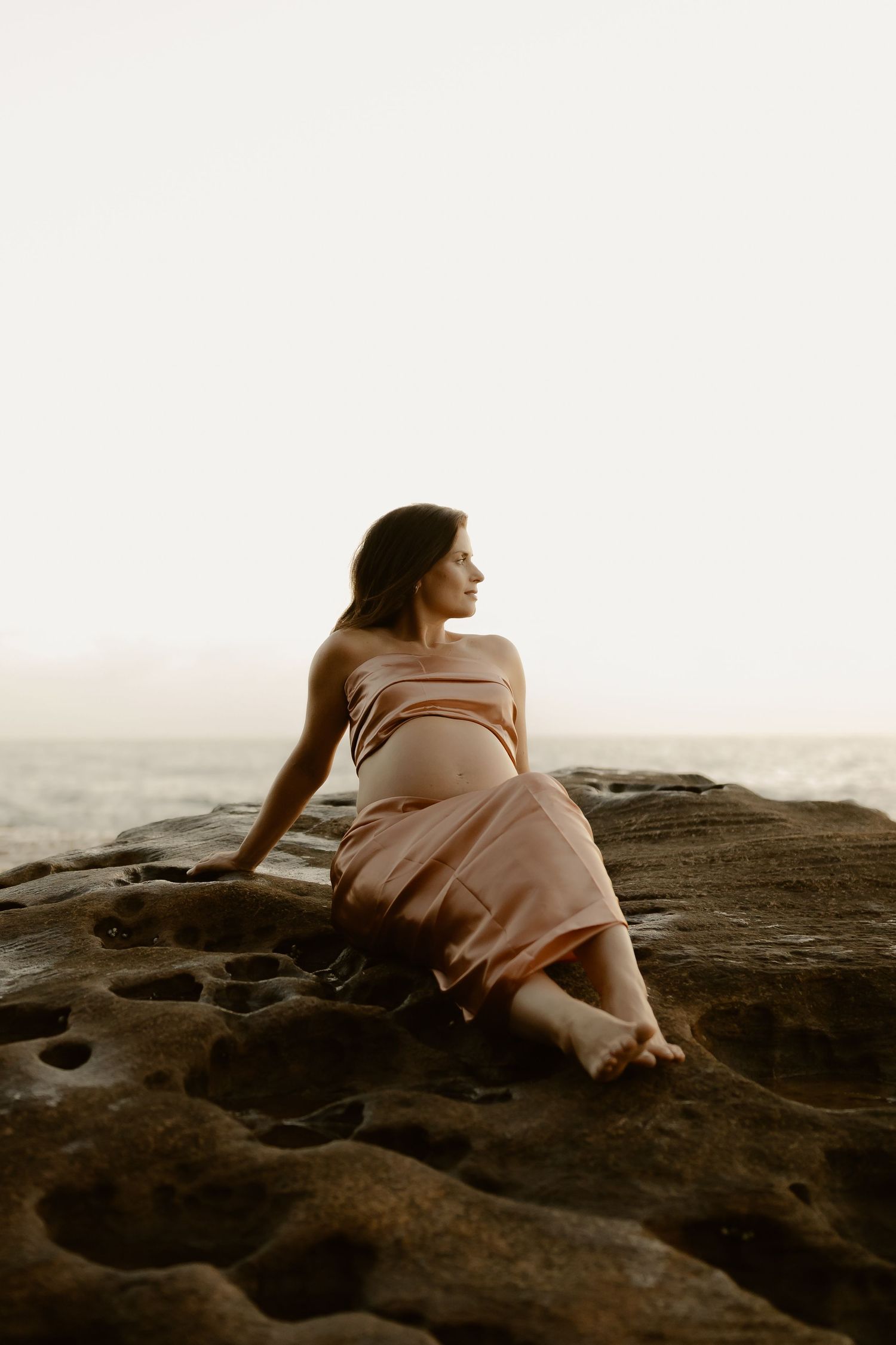A person in a flowing dress reclines on dark coastal rocks at sunset in a dreamy seaside photoshoot.