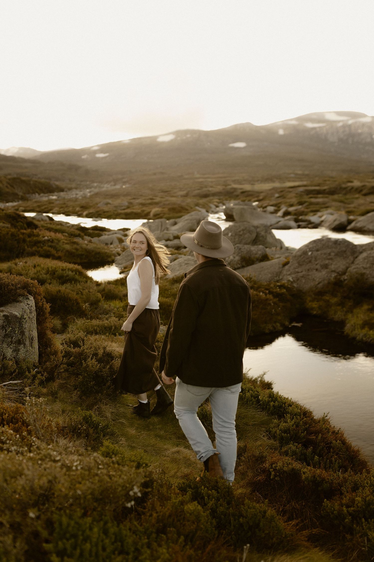 Two people walk hand in hand along a scenic mountain path with water and rolling hills in the background.