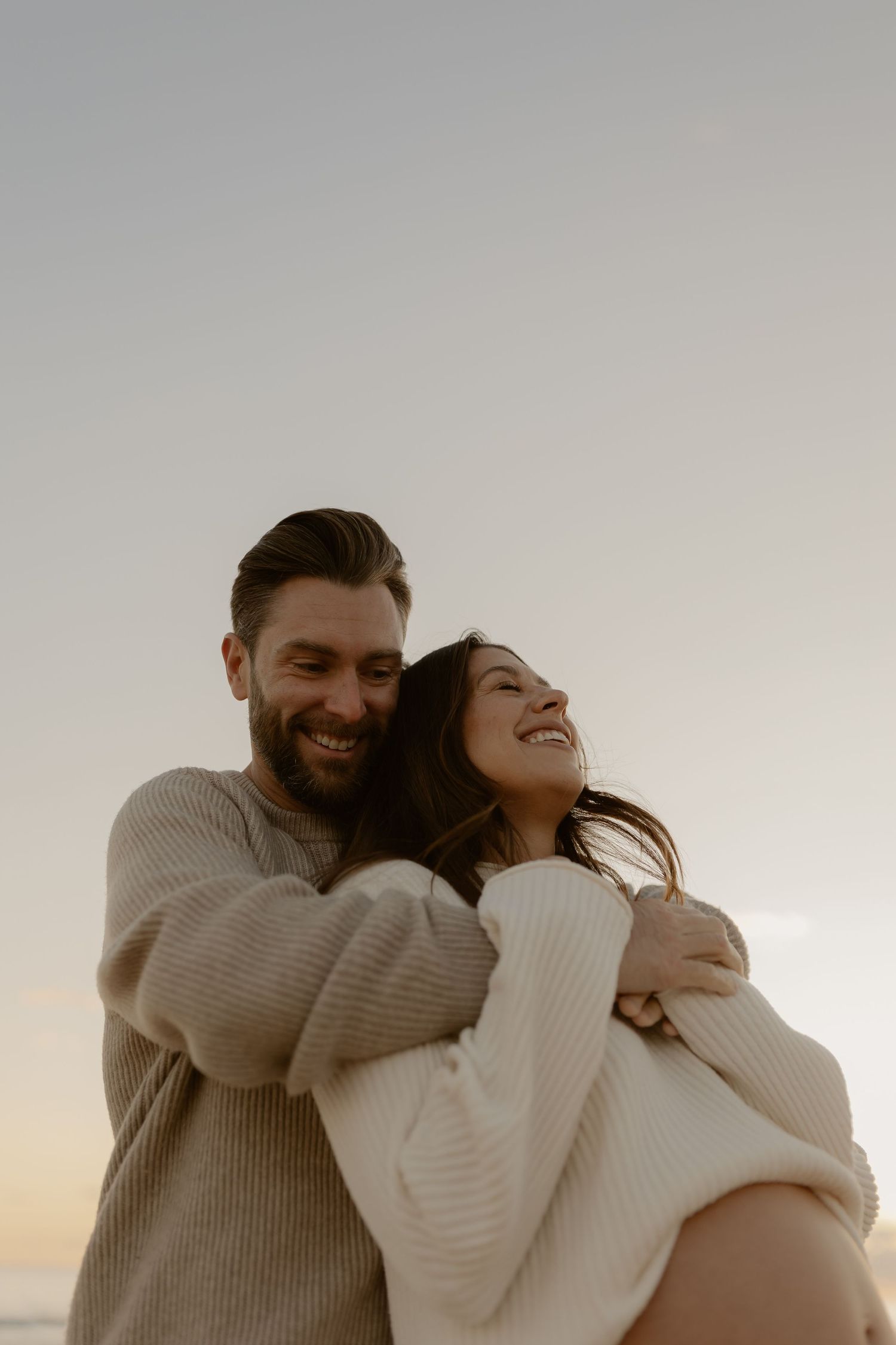 Couple in cozy sweaters sharing an intimate embrace against a bright background.