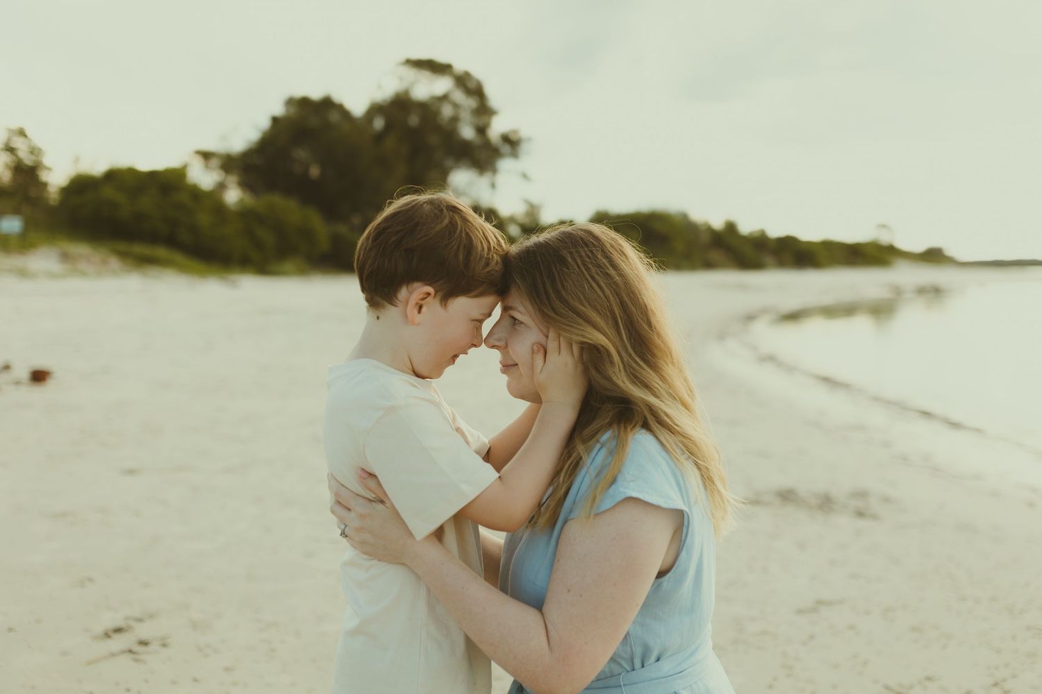 Romantic couple embracing on a scenic beach with water and trees in background.