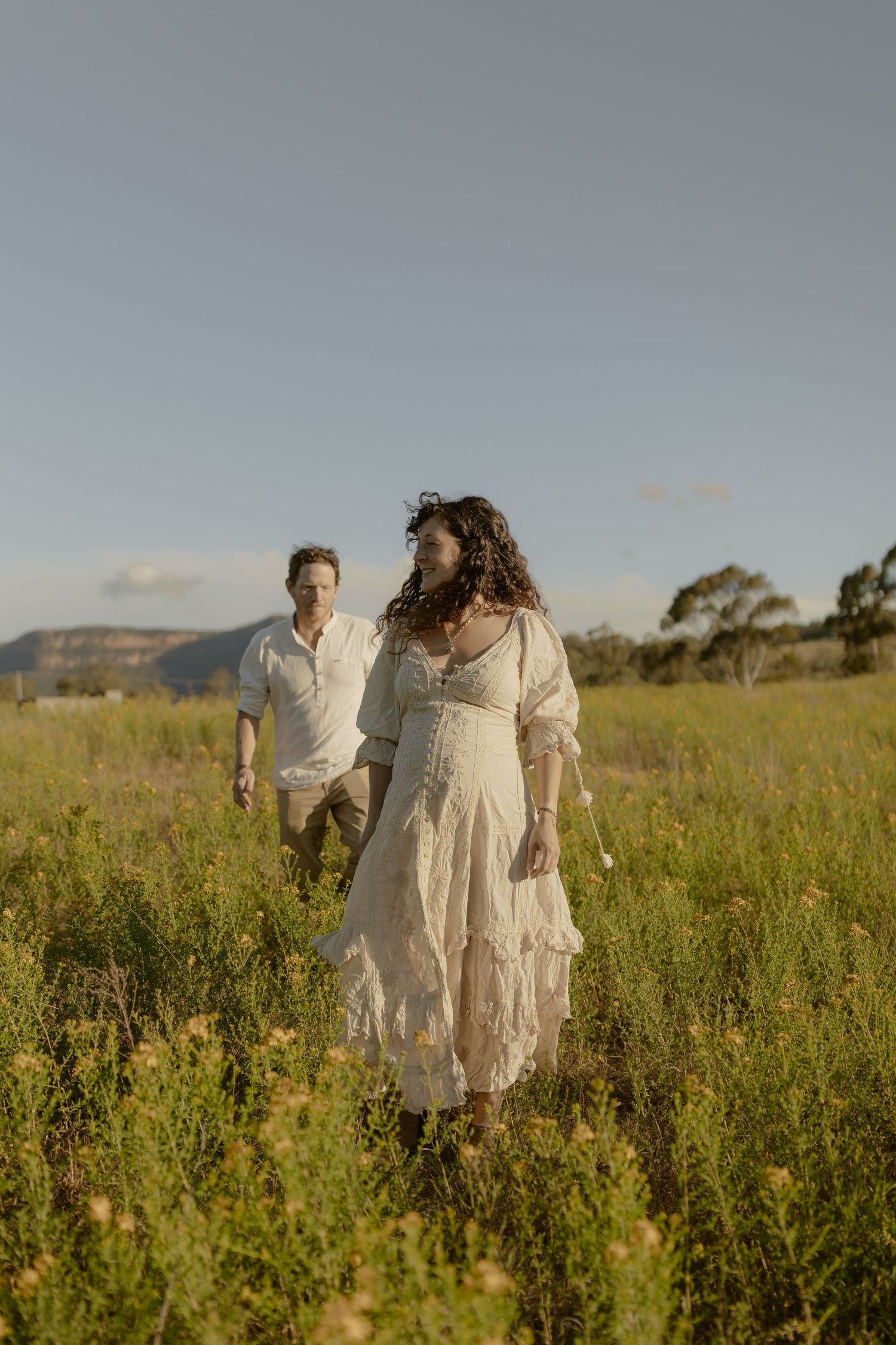 A couple in white dresses walks through a sunlit meadow of tall wildflowers with mountains in the background.