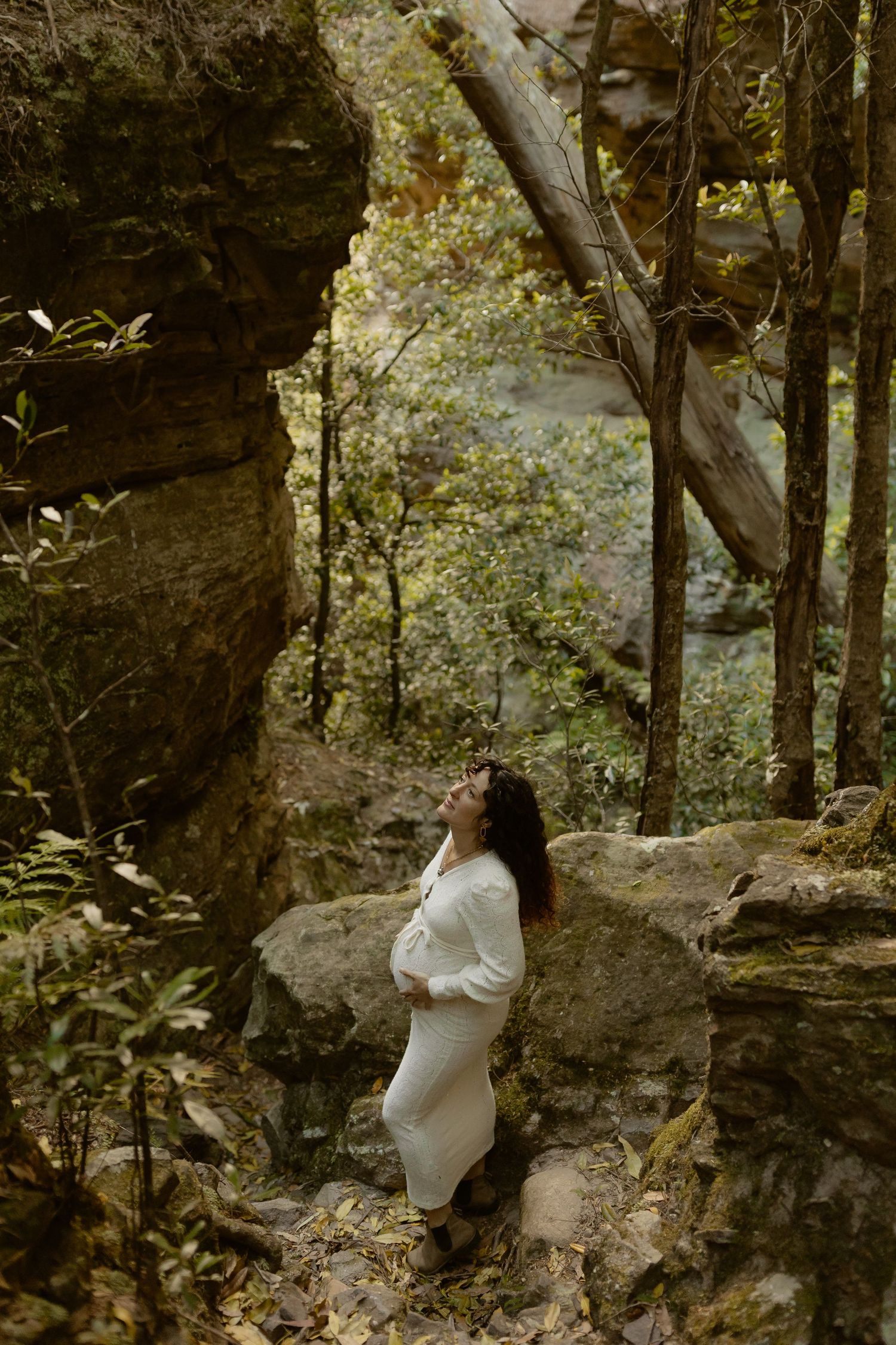 A figure in a white dress stands contemplatively among rocky cliffs and trees in a moody forest setting.