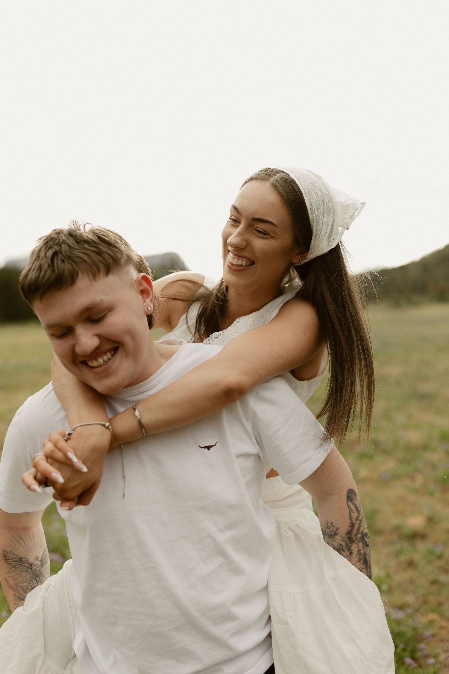 A playful couple in white clothing share a lighthearted moment outdoors.