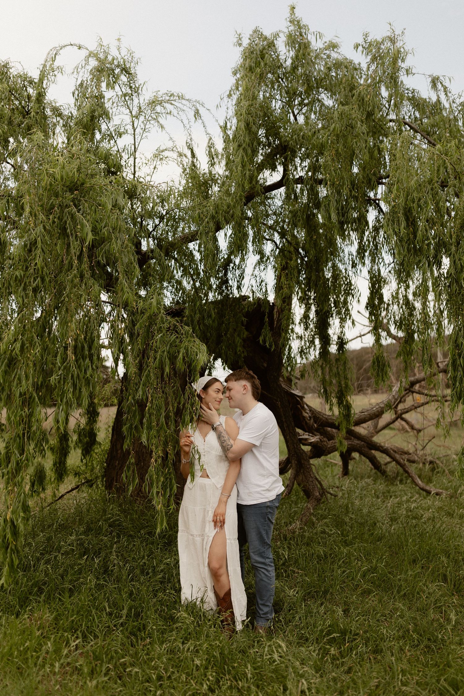 A couple shares a romantic moment under a drooping willow tree in a natural outdoor setting.