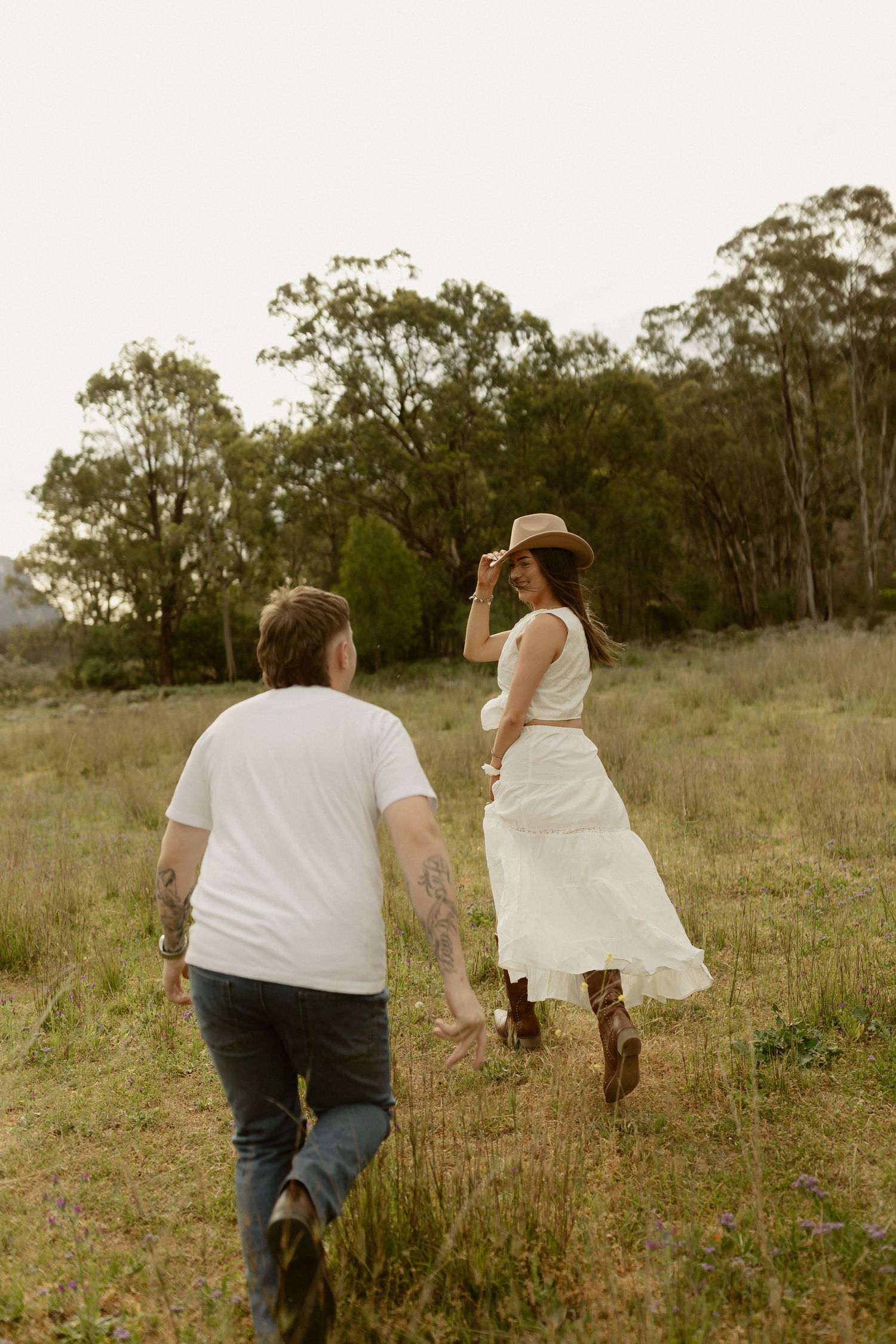 A couple runs playfully through a grassy field with trees in the background at sunset.