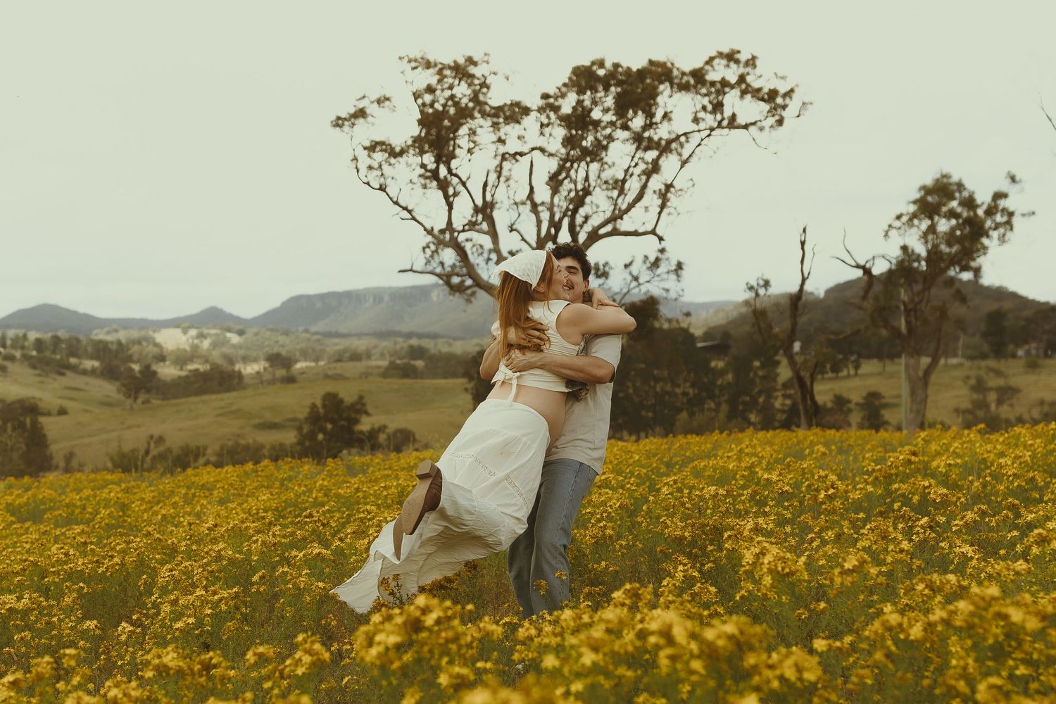 Two people walk hand in hand through a golden meadow with mountains visible in the distance.