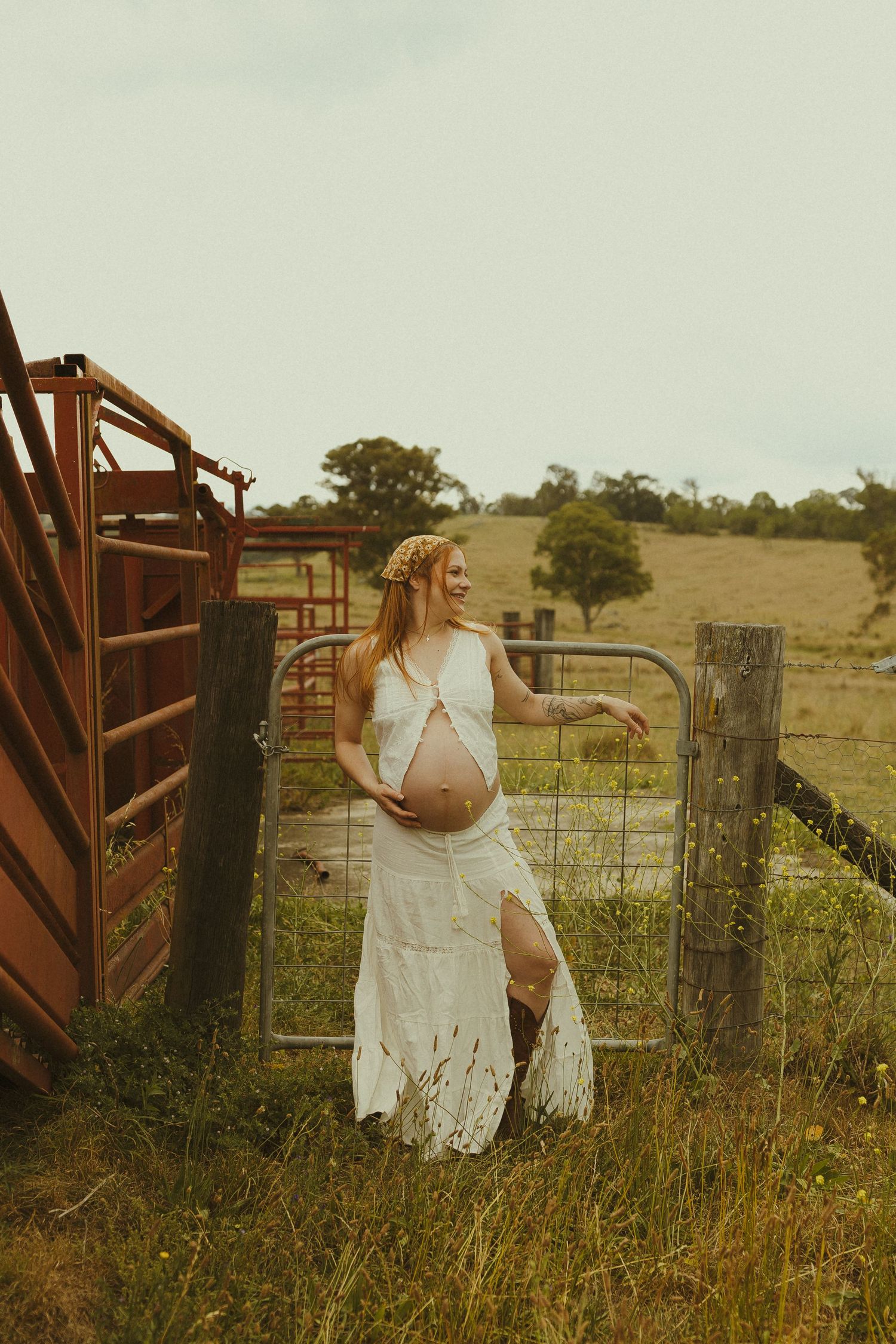 A rustic countryside photoshoot featuring a white lace dress against a metal farm gate and rolling hills.