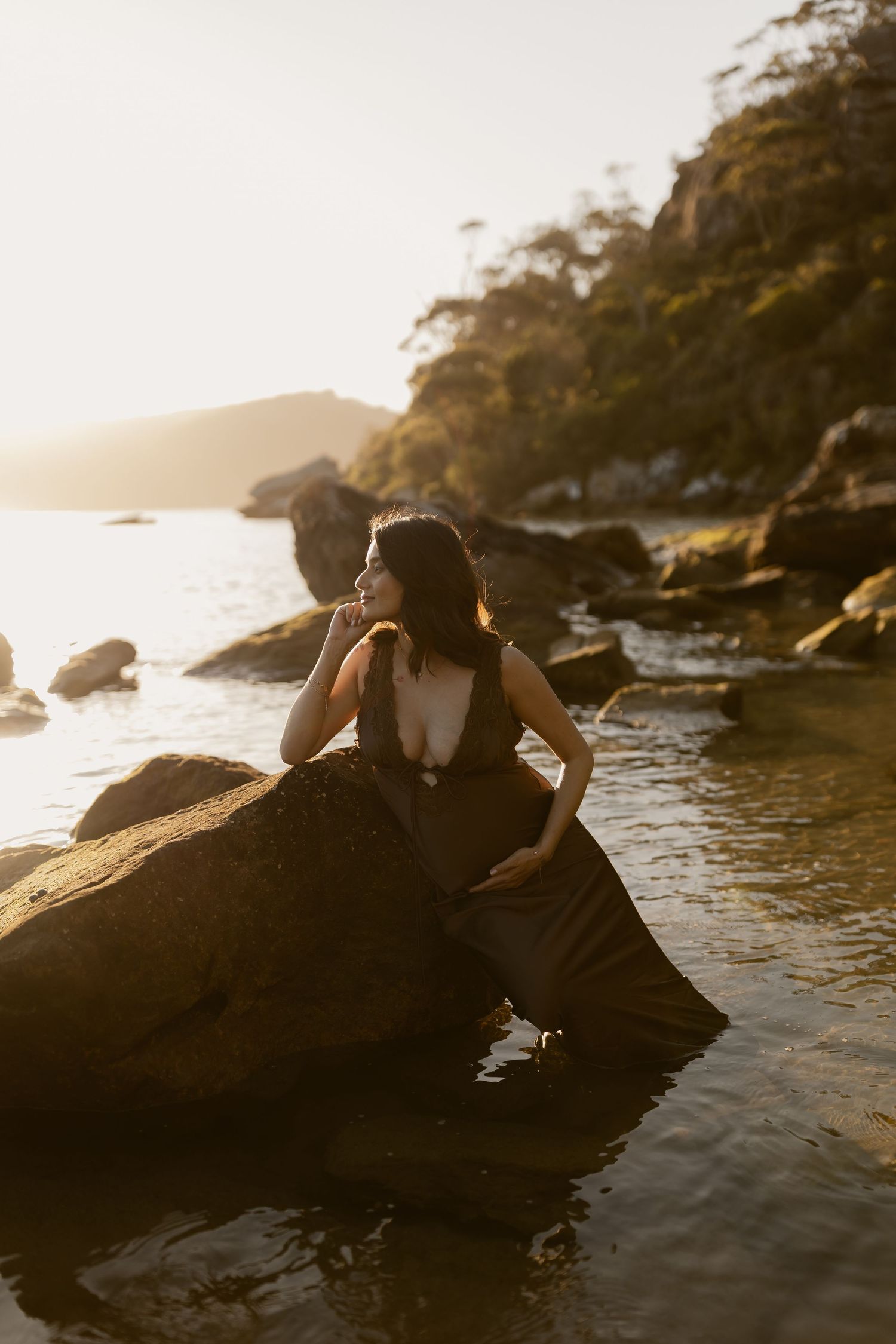 A romantic couple in formal attire poses intimately on coastal rocks during golden hour sunset by the ocean.