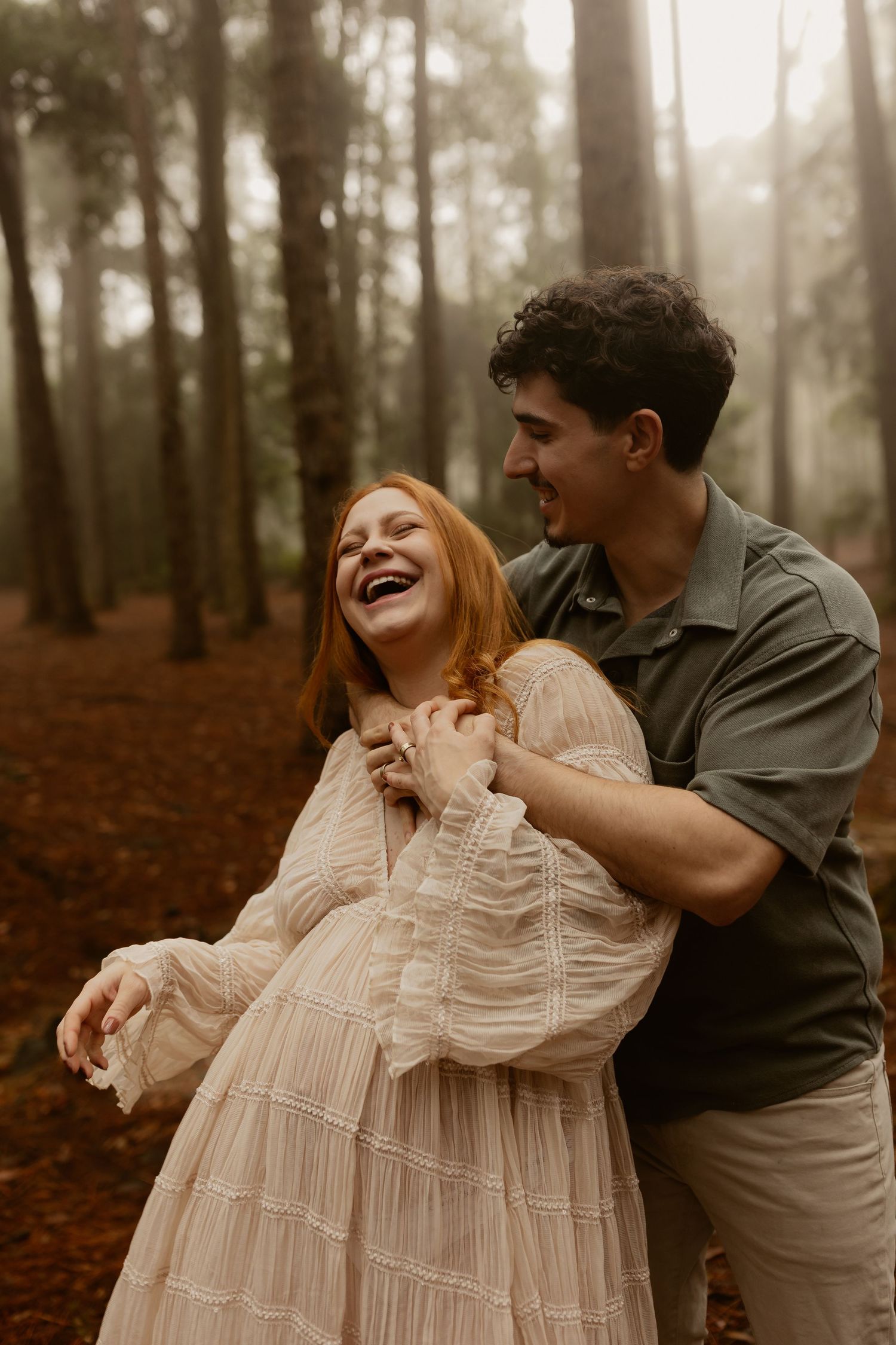 A couple shares a joyful moment together in a misty forest setting, with the woman wearing a flowing cream dress.