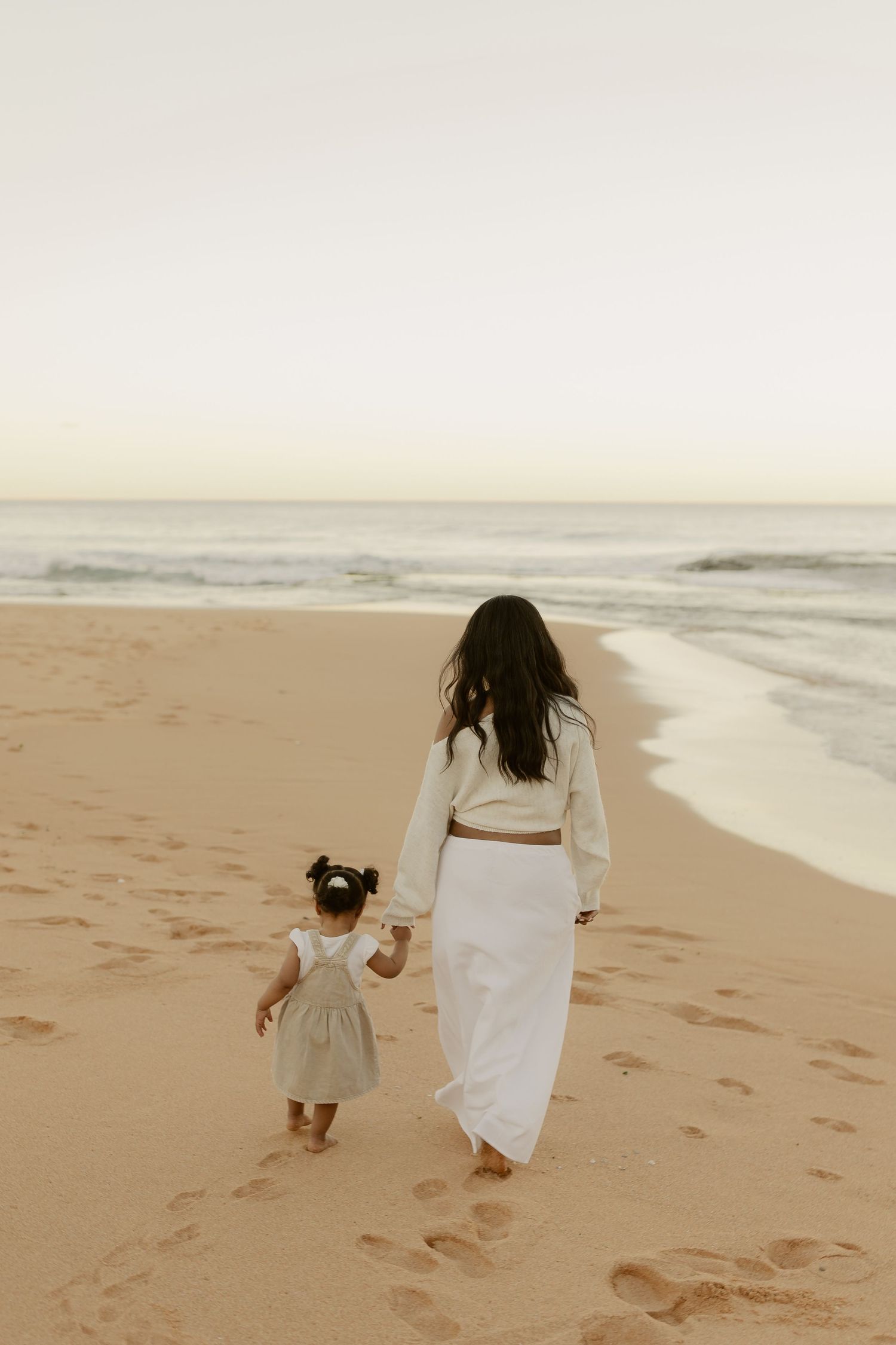 Mother and toddler daughter walking hand in hand toward ocean waves on sandy beach.