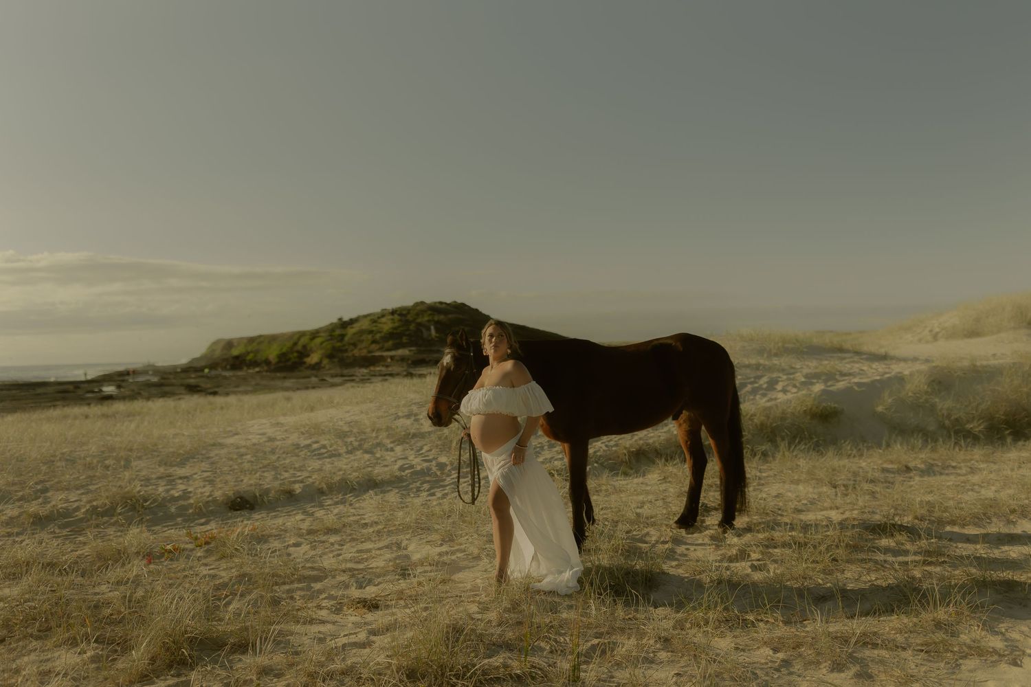 A woman in a white dress stands beside a dark horse on a sunlit coastal dune landscape.