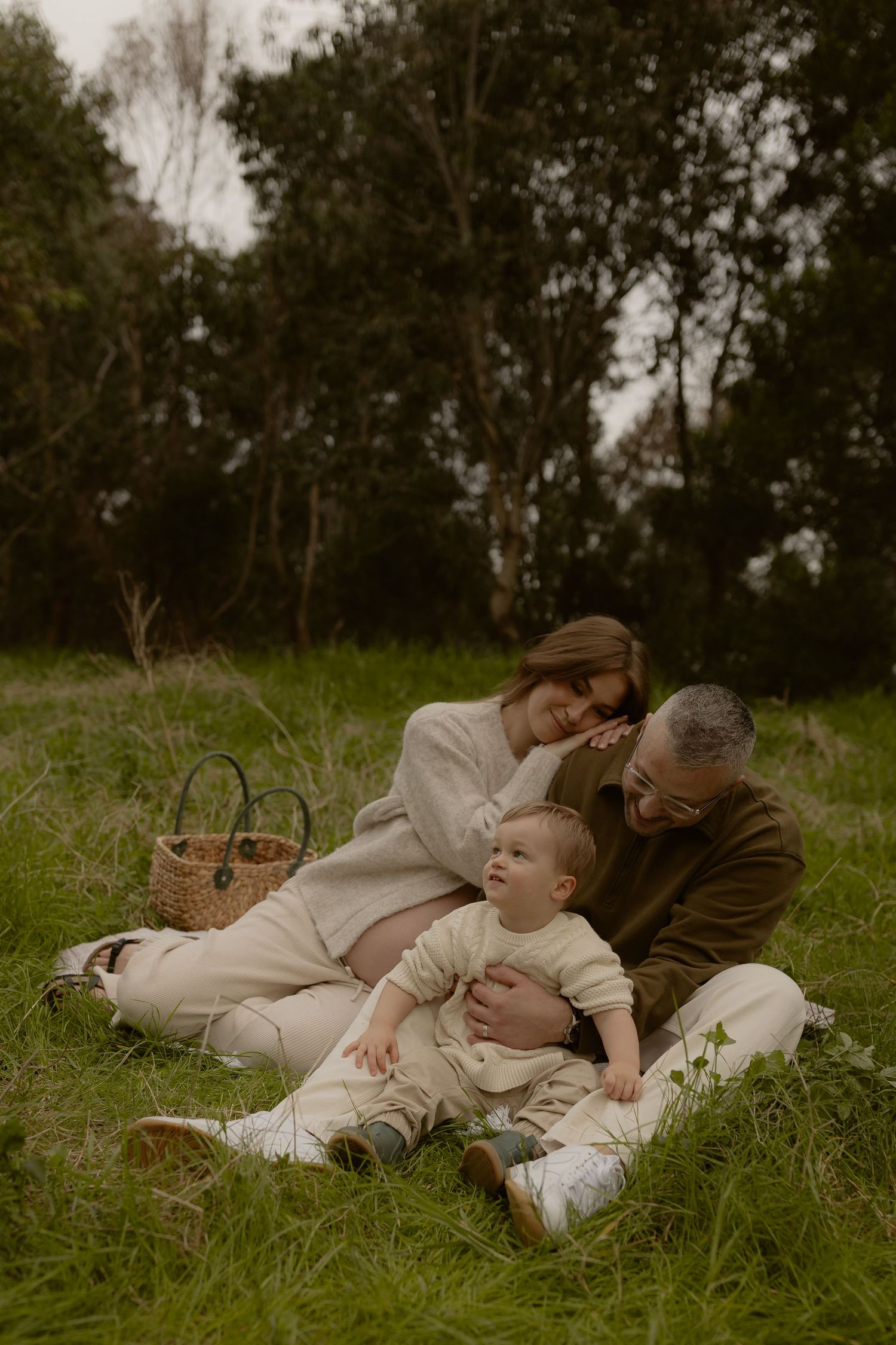Family sitting on grass in a park, parents lovingly gazing at their baby during a peaceful outdoor picnic.