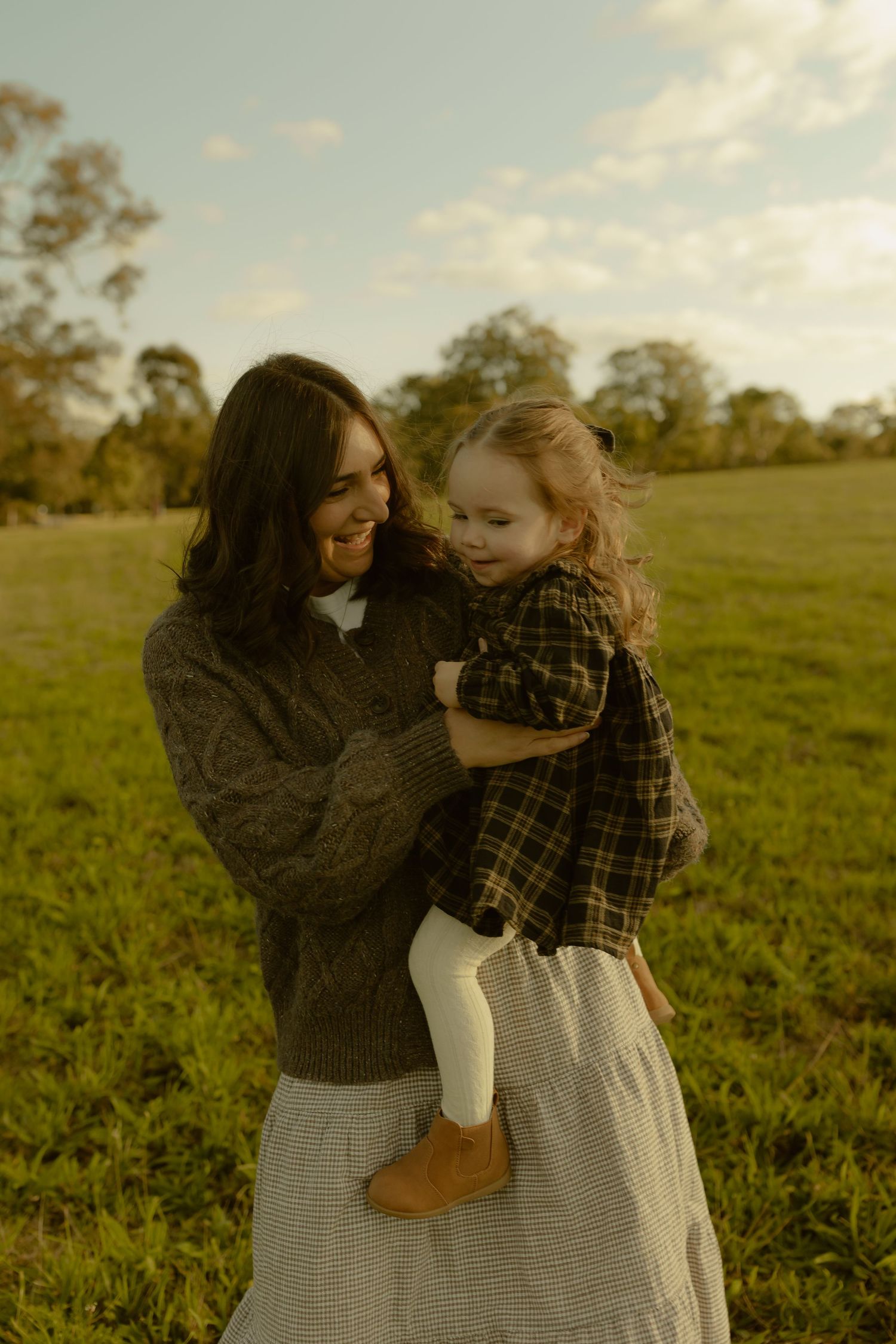 A smiling mother in a knit sweater holds her toddler daughter in a plaid shirt outdoors in a green field.