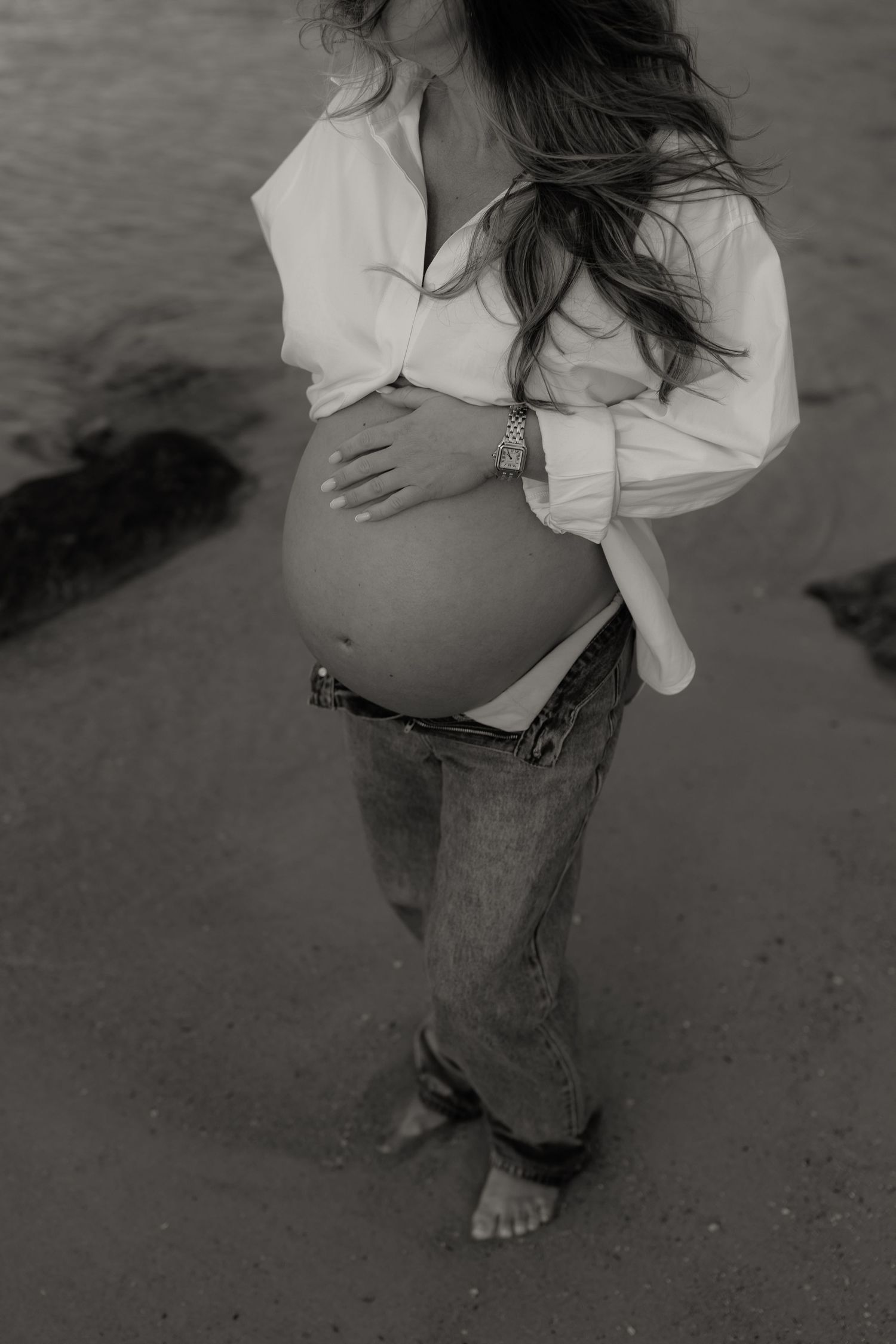 Black and white photo of a pregnant woman cradling her bare baby bump, wearing an open white shirt and jeans on a beach.