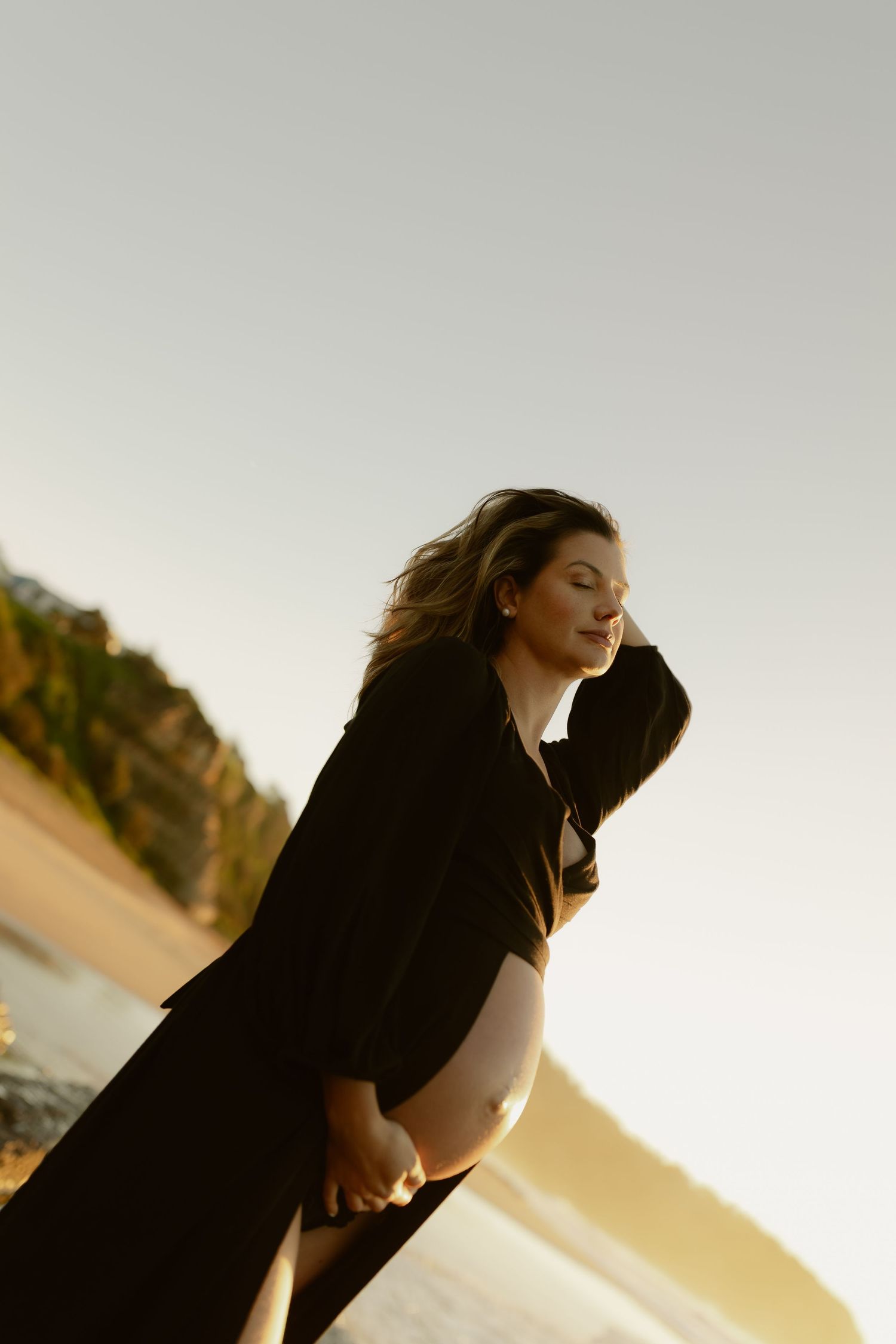 Pregnant woman in black robe cradling bare baby bump on golden beach at sunset.